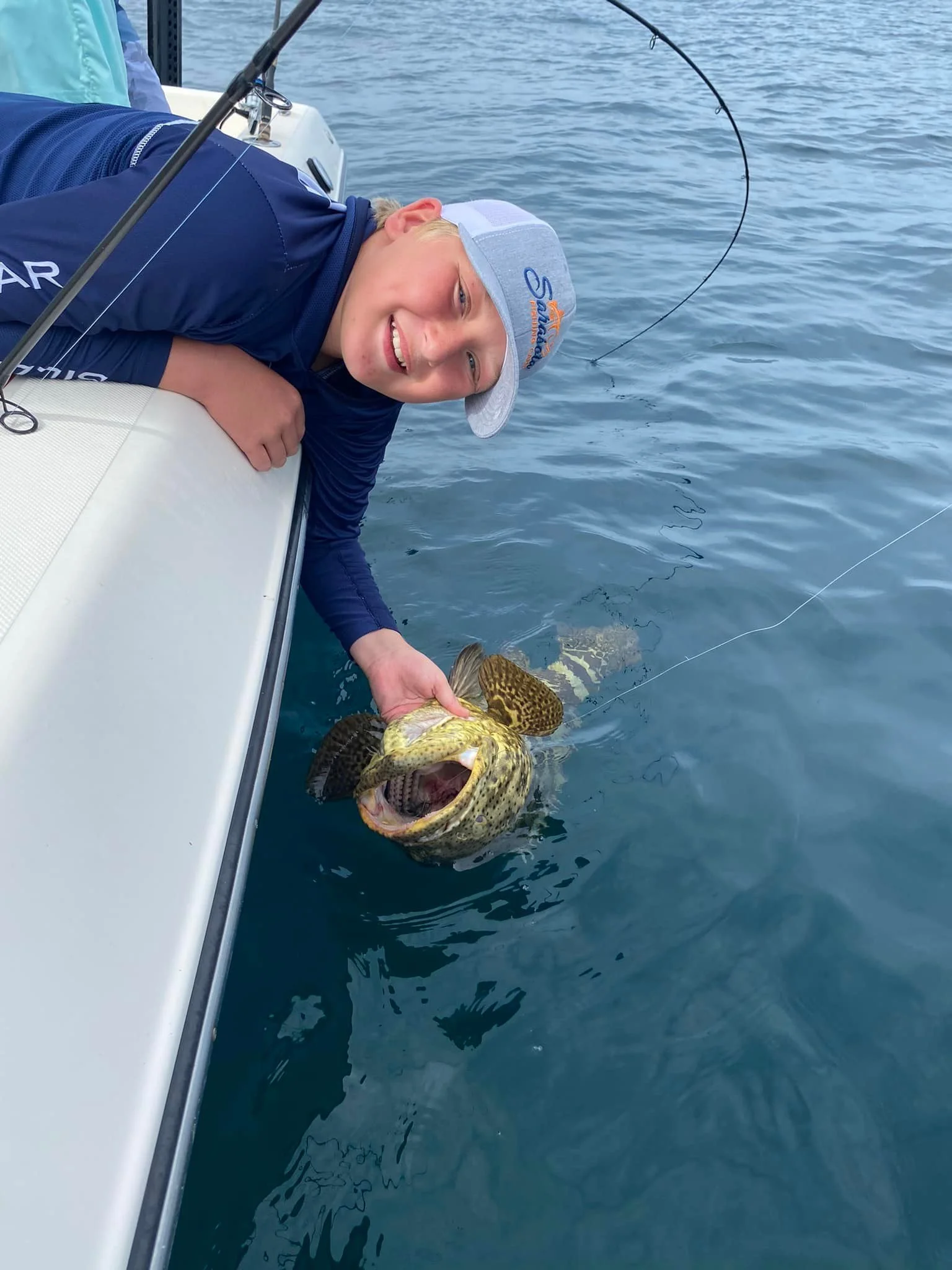A young person smiling while holding a large, spotted fish with wide open mouth, on a boat in the water.