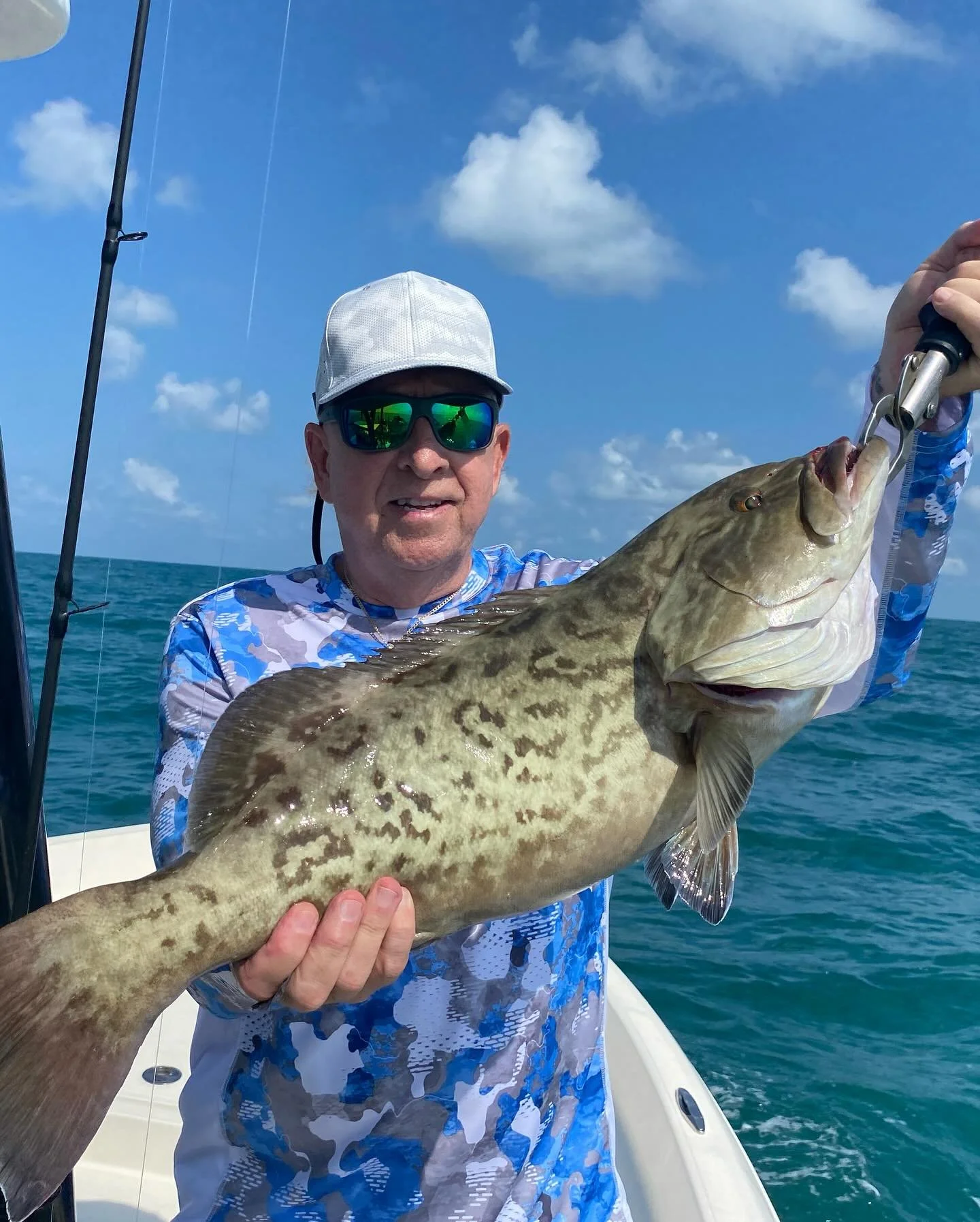 Man wearing sunglasses and a white cap holding a large, spotted fish on a boat in the ocean under a partly cloudy sky.