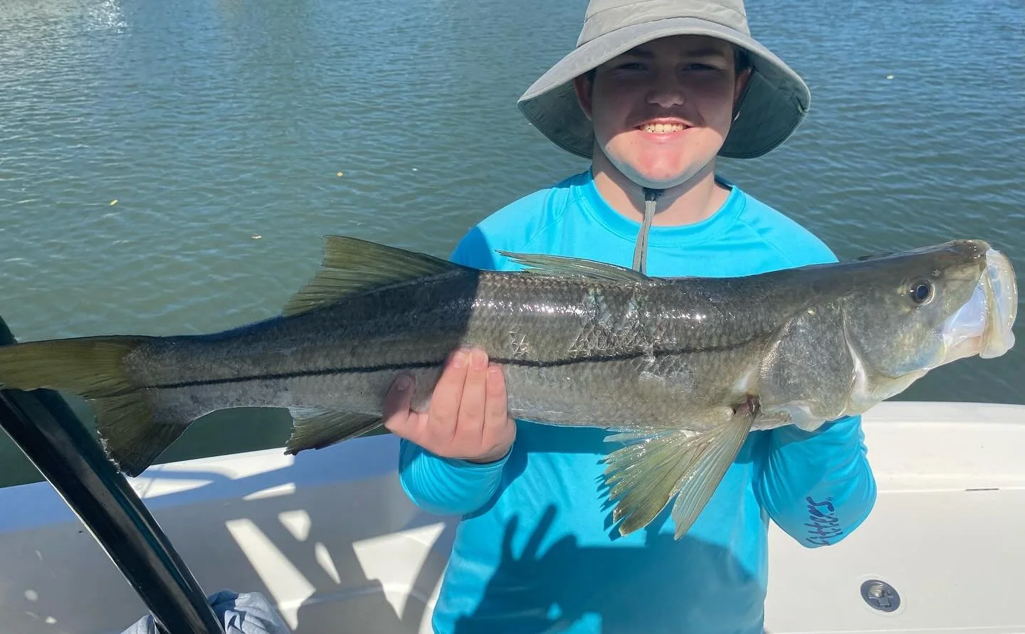 A boy in a blue shirt and wide-brimmed hat holding a large fish on a boat with water in the background.