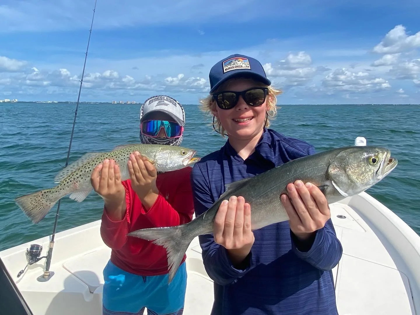 Two people on a boat holding caught fish, with a person in the front holding a large gray fish and another person in the back holding a smaller fish, both wearing sunglasses and hats, with an ocean and cloudy sky in the background.