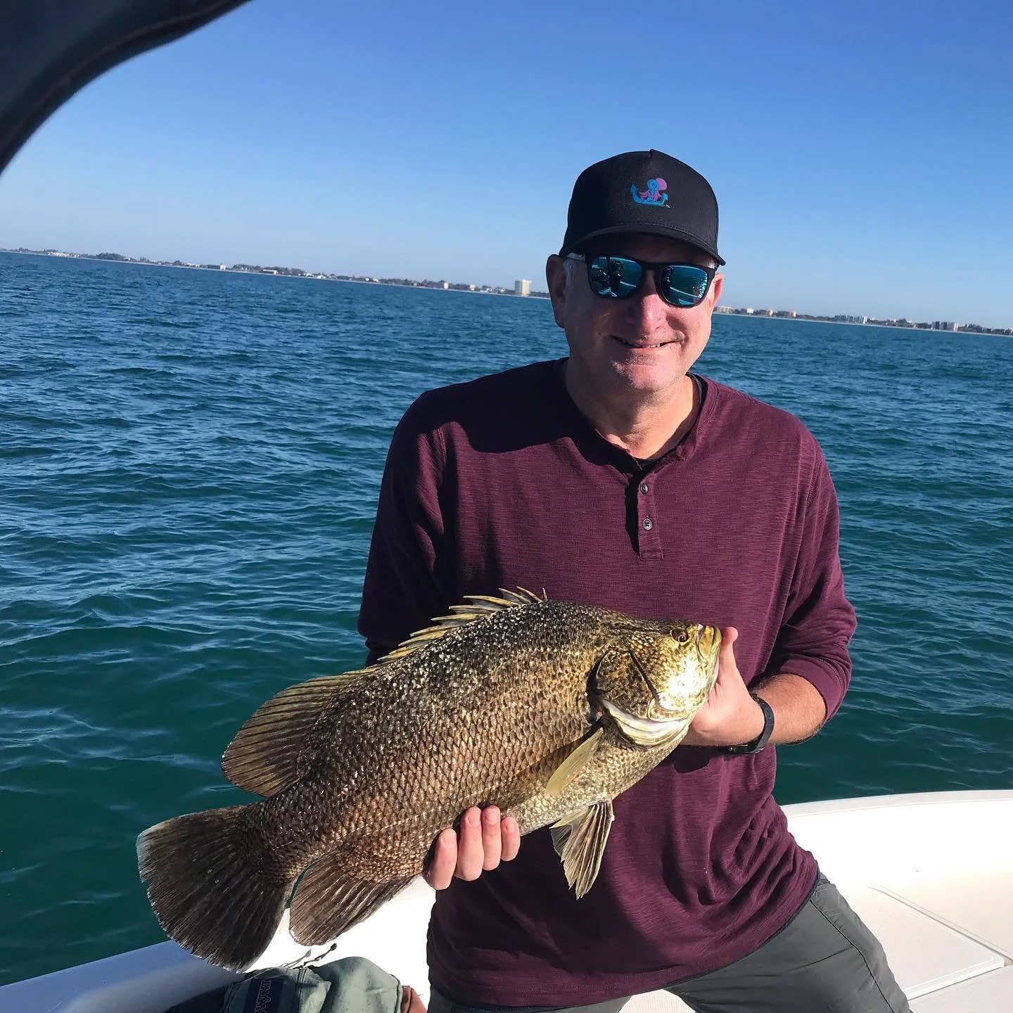 Man holding a large fish on a boat with water and city skyline in the background