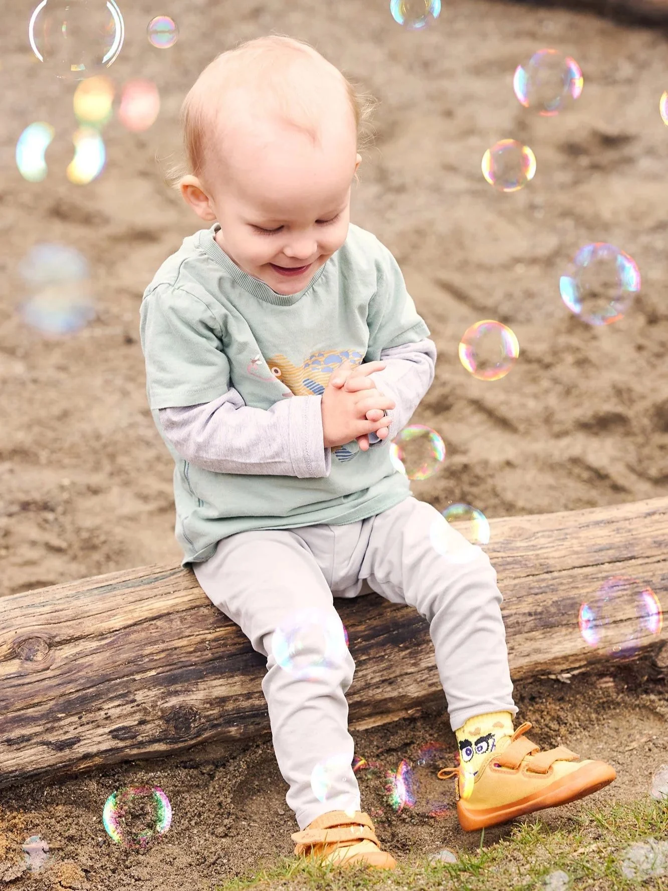 Ein lachendes kleines Kind sitzt auf einem Baumstamm im Freien, umgeben von schimmernden Seifenblasen, mit Sandboden im Hintergrund.