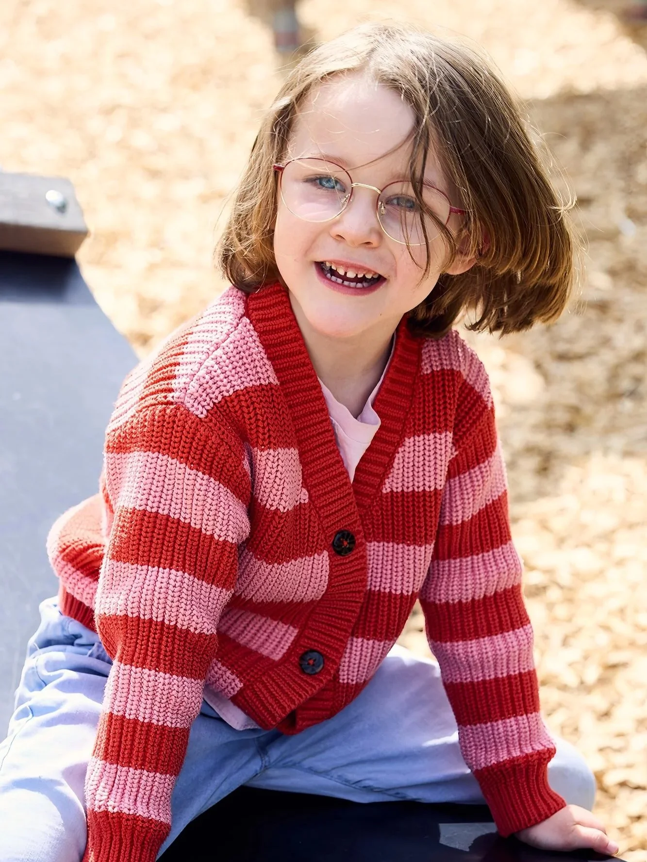 Junges, lächelndes Kind mit Brille trägt rot-rosa gestreiften Pullover, sitzt draußen auf einer Schwingmatte.