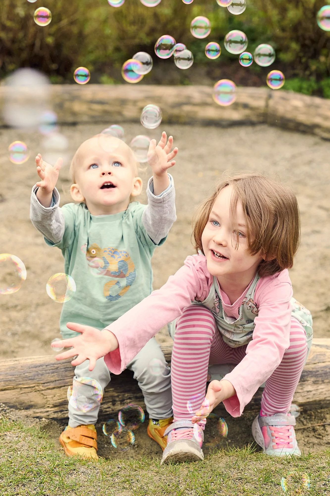 Zwei Kinder, ein Junge und ein Mädchen, spielen draußen auf einer Wiese mit Seifenblasen. 