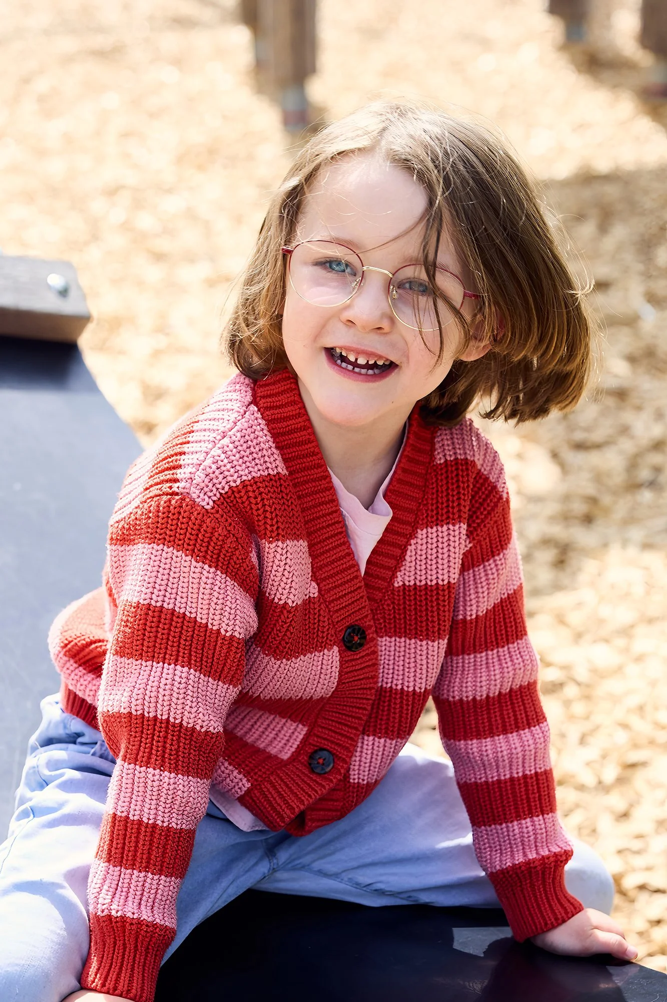 Junges, lächelndes Kind mit Brille trägt rot-rosa gestreiften Pullover, sitzt draußen auf einer Schwingmatte.