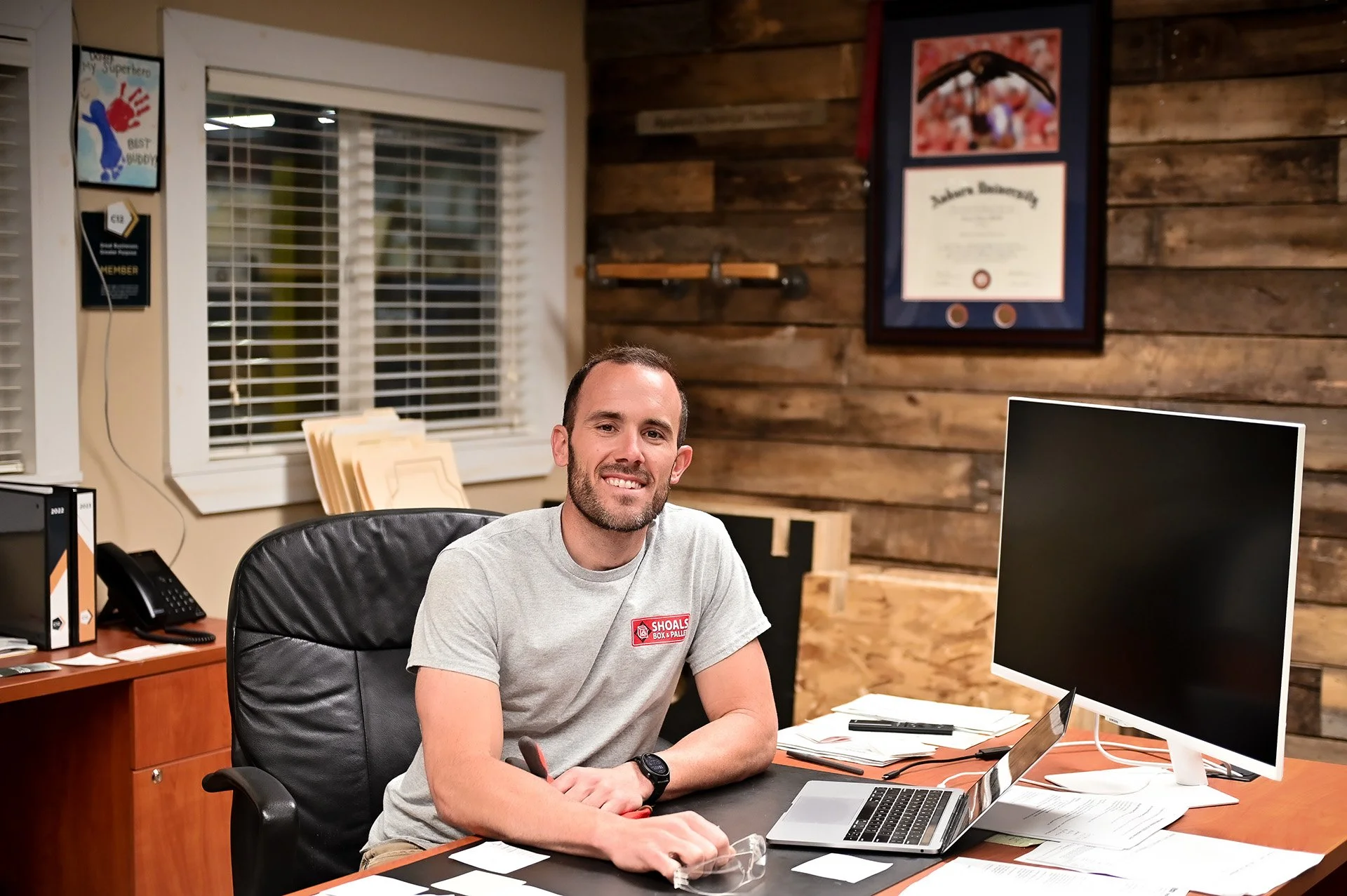 A man sits at a desk in an office, smiling, with computer equipment, papers, and a framed certificate on the wooden wall behind him.