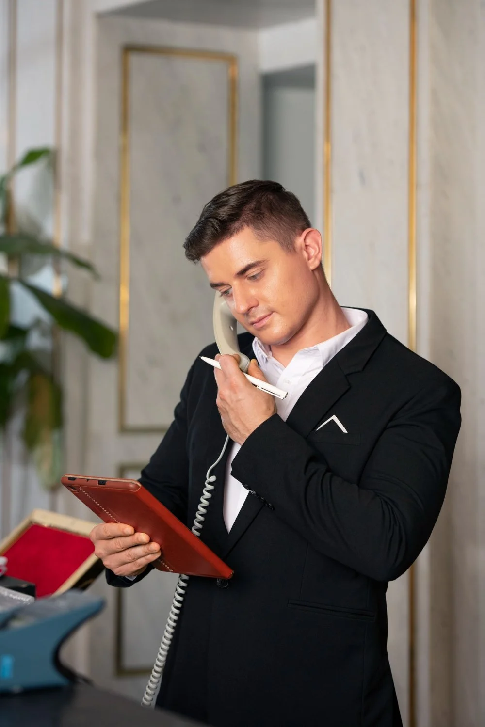 Businessman in black suit talking on landline phone while looking at a red notepad in an elegant office.