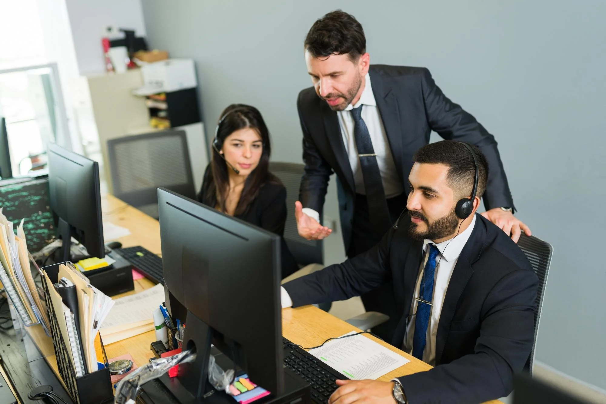Three colleagues, two men and one woman, in a modern office, working at desks with computer monitors, discussing work with the man standing and gesturing.