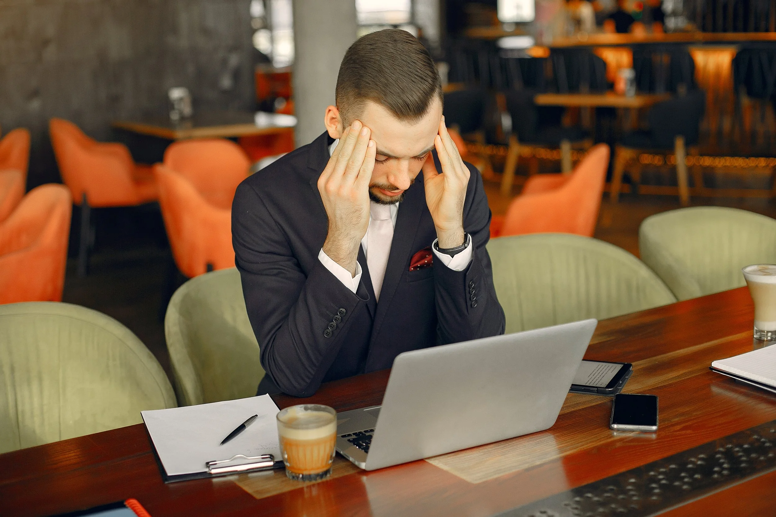 A man in a dark suit sitting at a wooden table in a cafe or restaurant, holding his head with both hands, looking down at a laptop. There are a notebook, pen, smartphone, and a glass of coffee on the table.