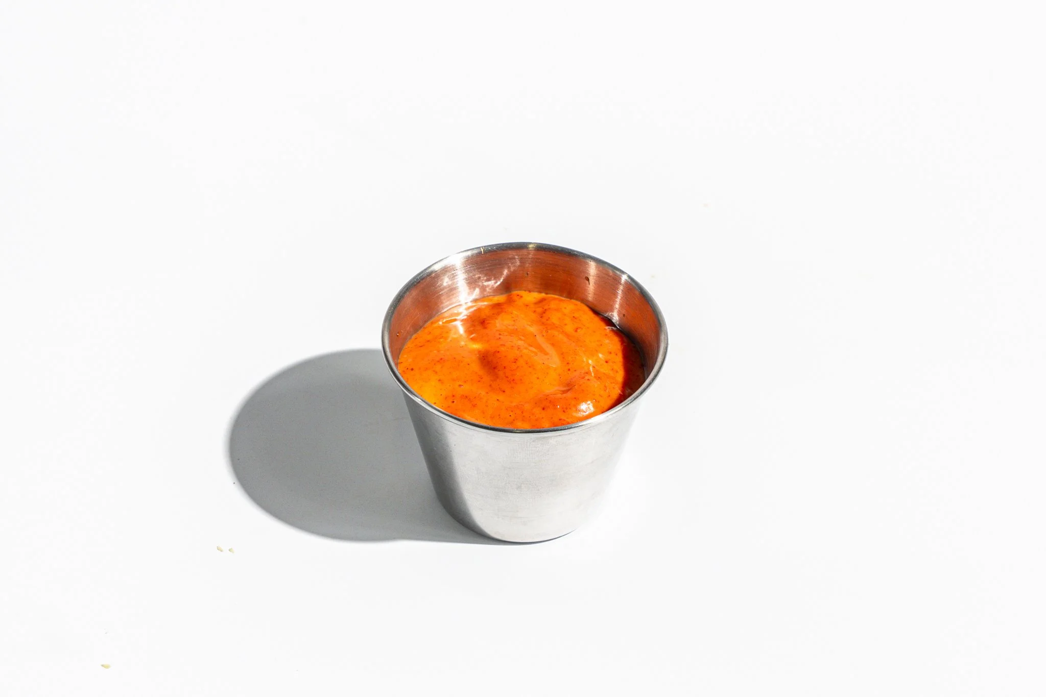 A stainless steel cup filled with orange chili sauce on a white surface with a shadow.
