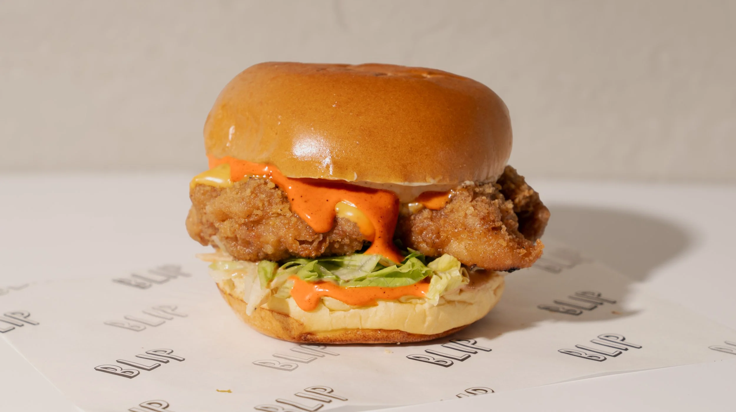 Fried chicken sandwich with lettuce, sauce, and bun on a white paper with black text, on a white surface.