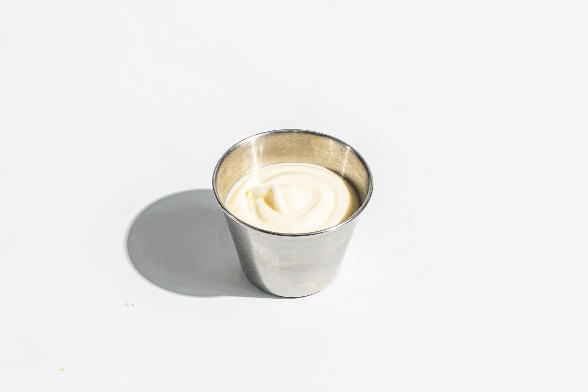 A metal cup filled with mayonnaise casting a shadow on a white surface.