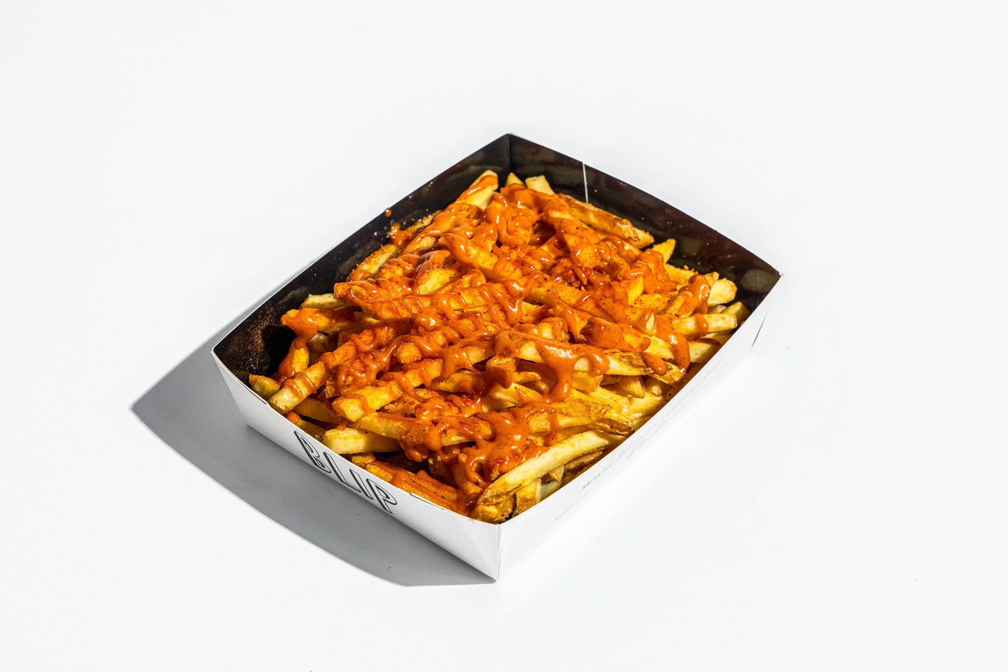 A rectangular cardboard container of French fries topped with cheese and spicy sauce on a white surface.