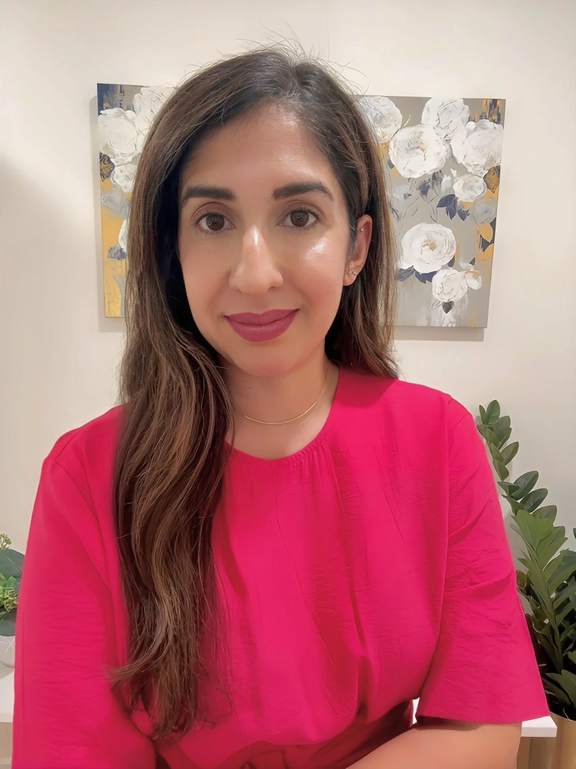 A woman with brown hair and light makeup wearing a bright pink top in front of abstract floral artwork and indoor plants.