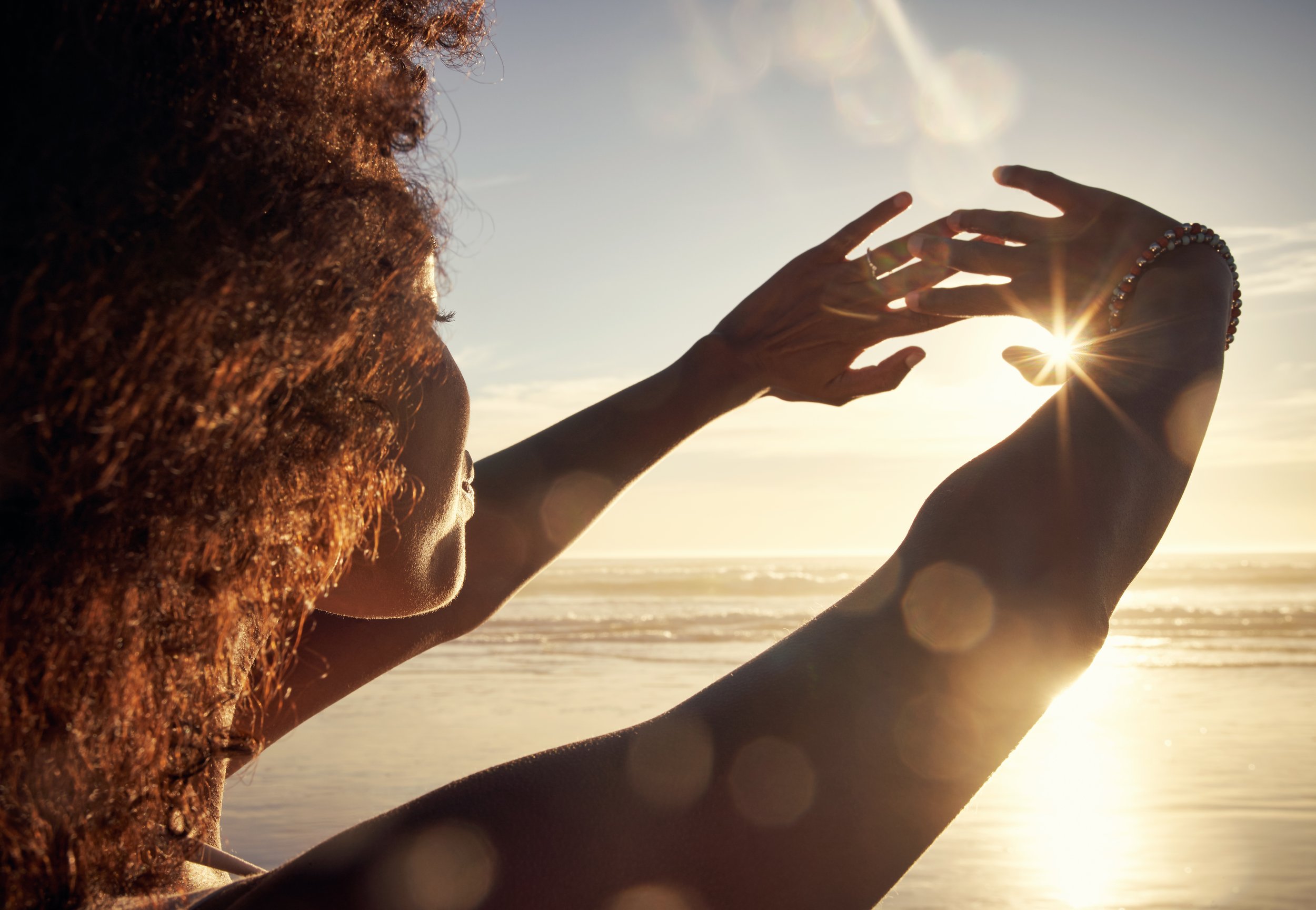A woman with curly hair holding a shoe up to the sun on a beach at sunset, creating a starburst effect.