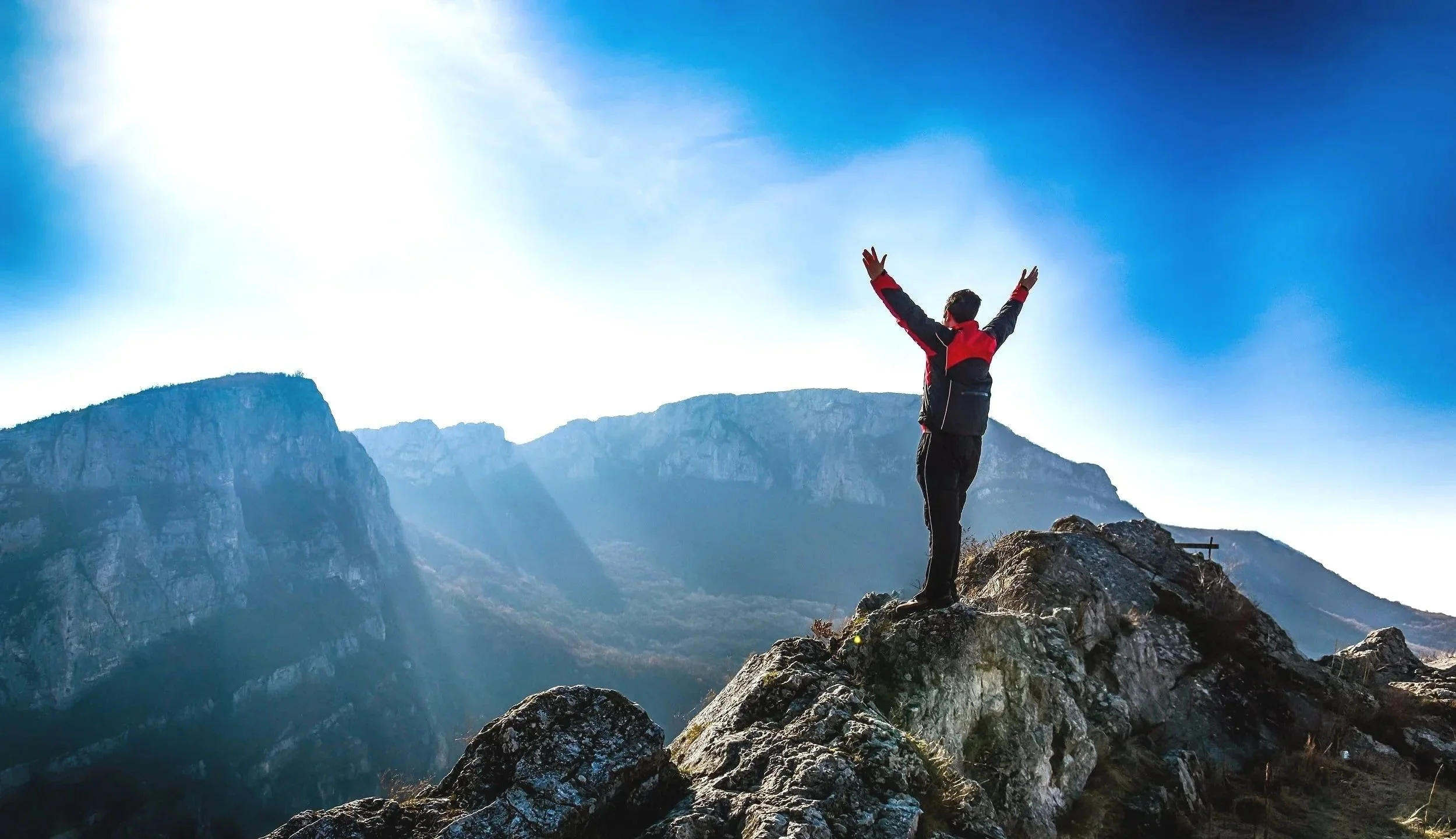 A person standing on a rocky mountain peak with arms raised in victory, facing a range of mountains under a clear, blue sky with sunlight streaming in.
