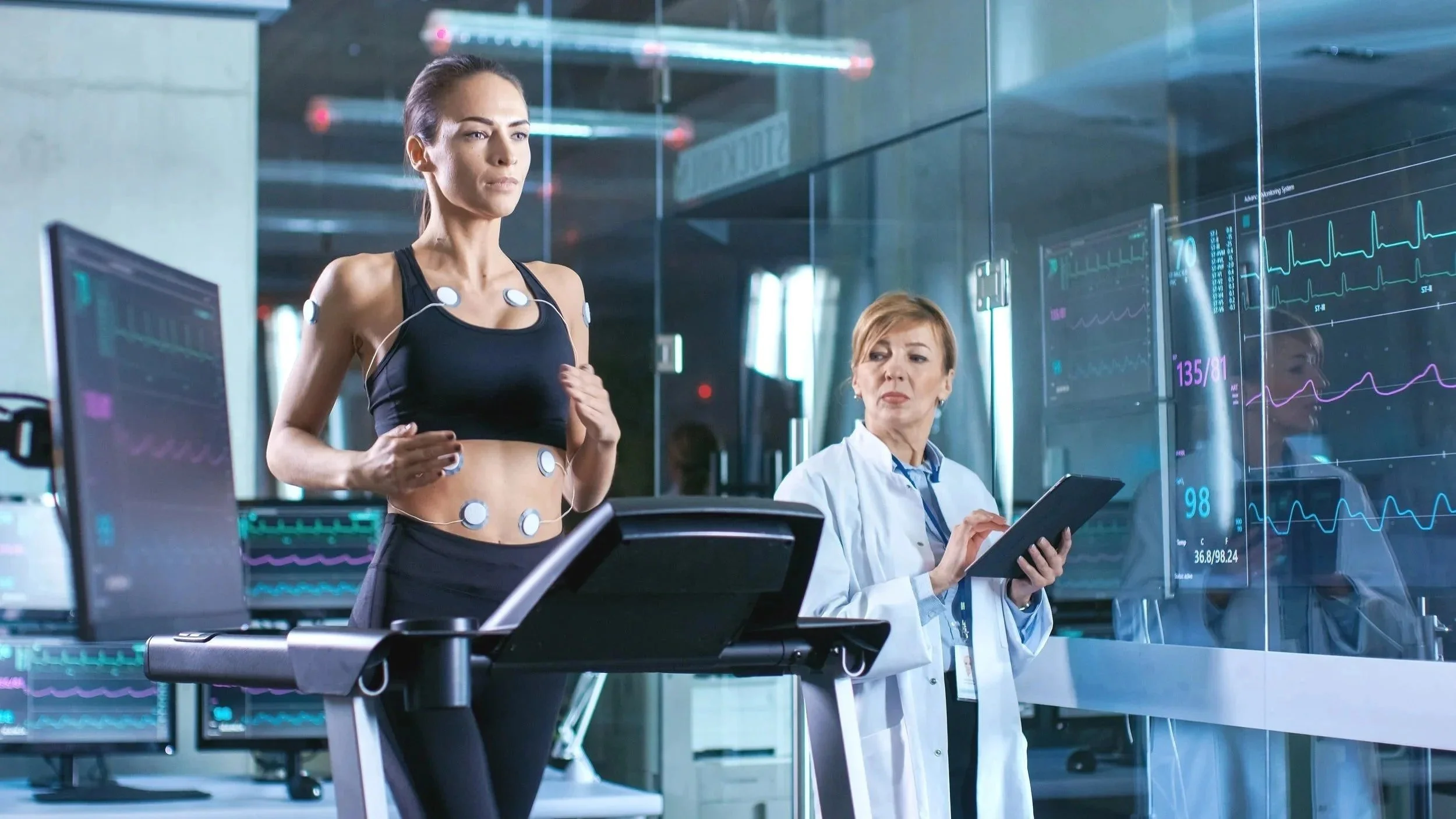 A woman undergoing a fitness test on a treadmill with electrodes attached to her body, monitored by a technician with a tablet, in a high-tech medical lab with multiple screens displaying heart rate data.