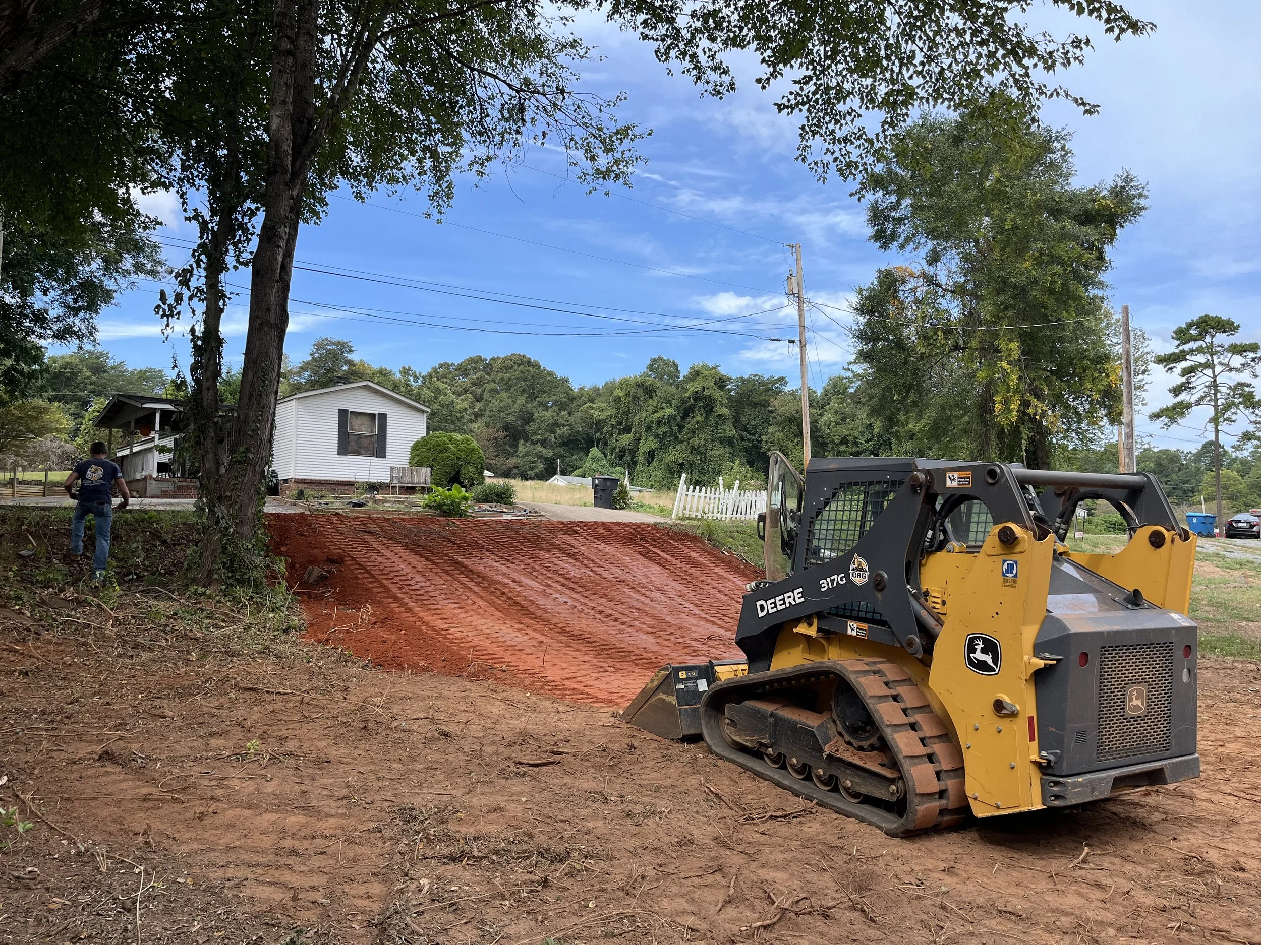 A small yellow and black John Deere compact track loader parked on a dirt surface in front of a house, with a person standing nearby, indicating construction or landscaping work.