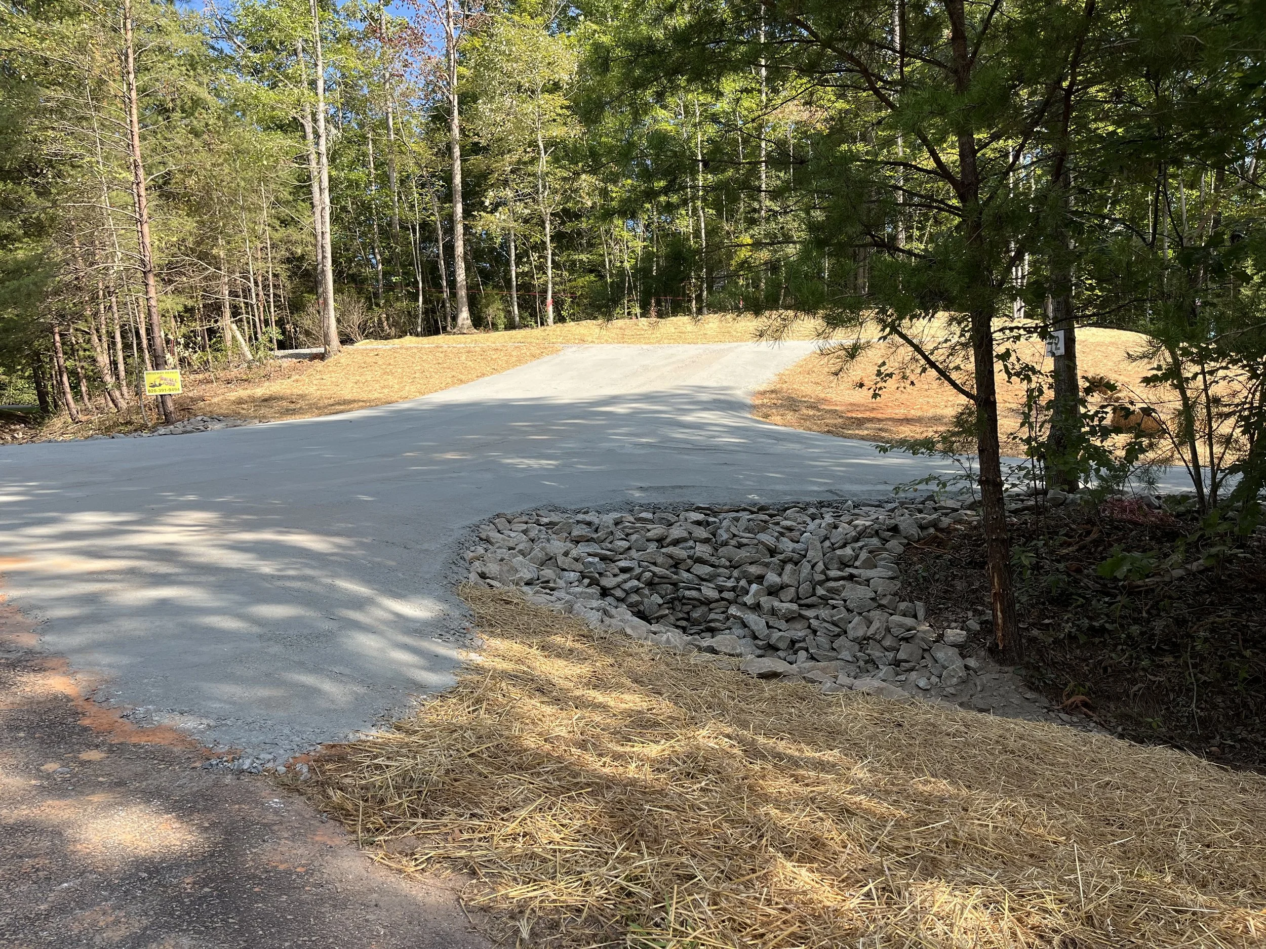 A winding gravel road under construction in a forested area, with trees on both sides and a pile of rocks near the edge of the road.