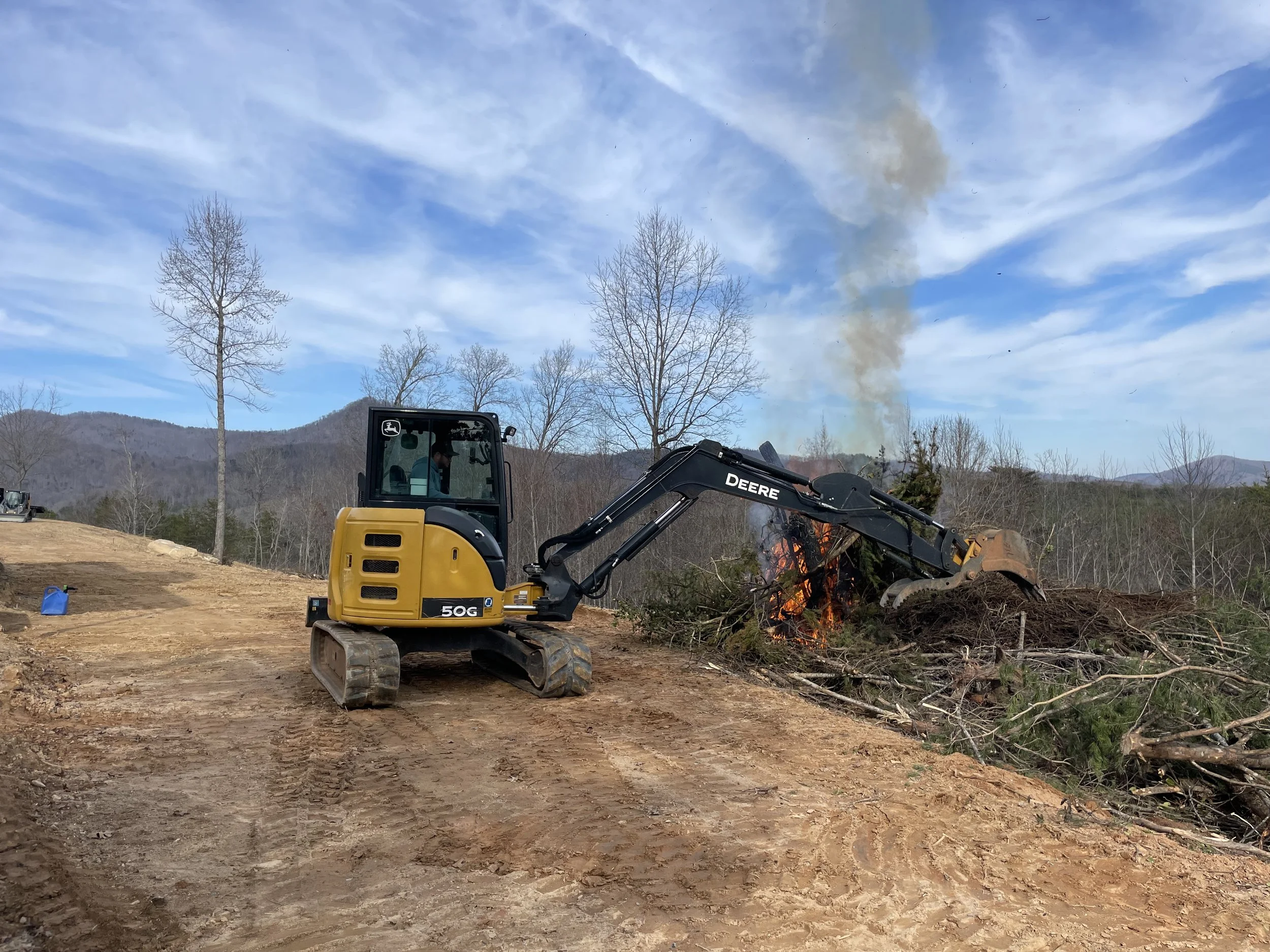 A small John Deere excavator demolishing a pile of fallen branches and brush on a dirt construction site outdoors, with mountains, leafless trees, and blue sky in the background.