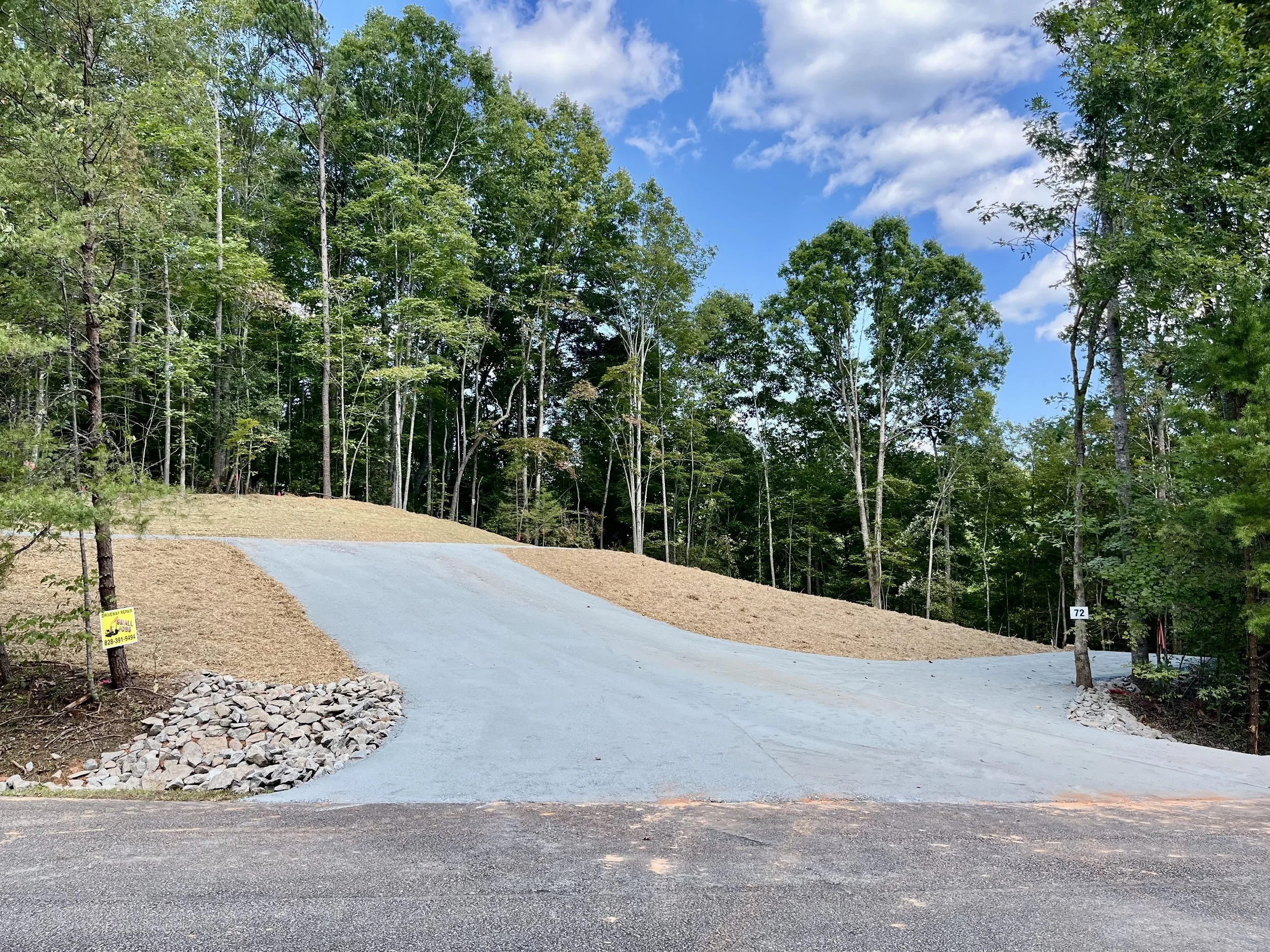 A newly paved driveway leading up a wooded hill, with trees on both sides and a cloudy sky overhead.