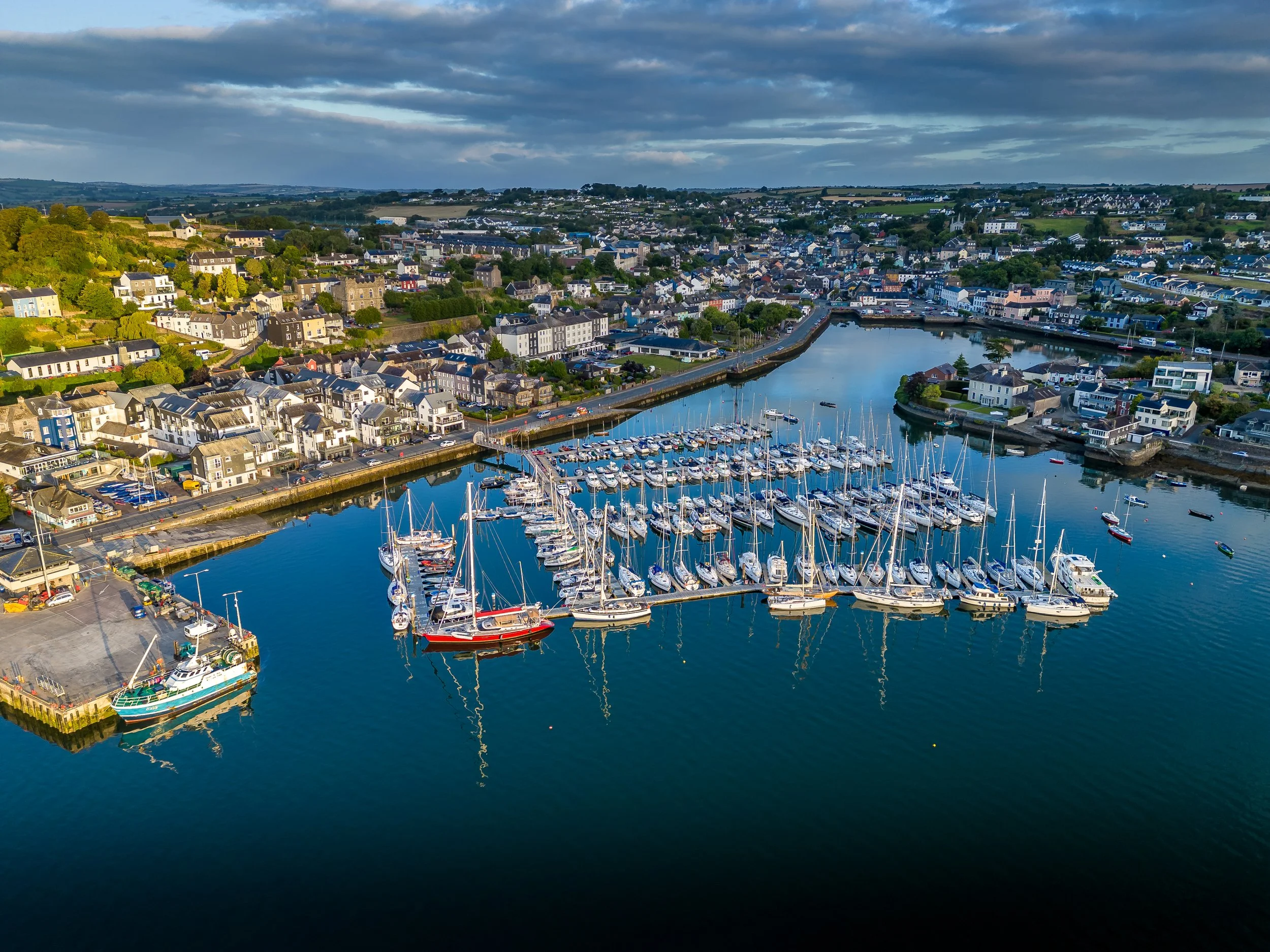 Aerial view of a marina with numerous docked sailboats and yachts, surrounded by a coastal town with houses and buildings, and hills in the background under a sky with scattered clouds.