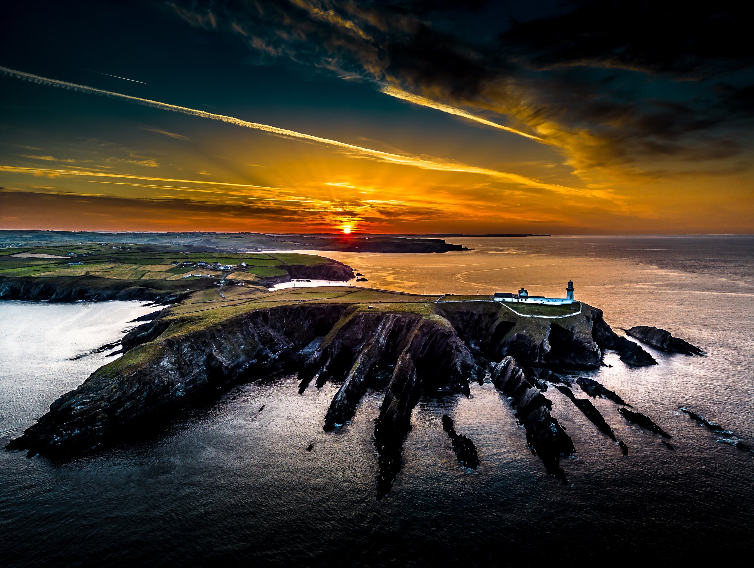 A coastal landscape at sunset showing a lighthouse on a rocky promontory, with green fields and small buildings, and a dramatic sky with clouds and contrails over the ocean.