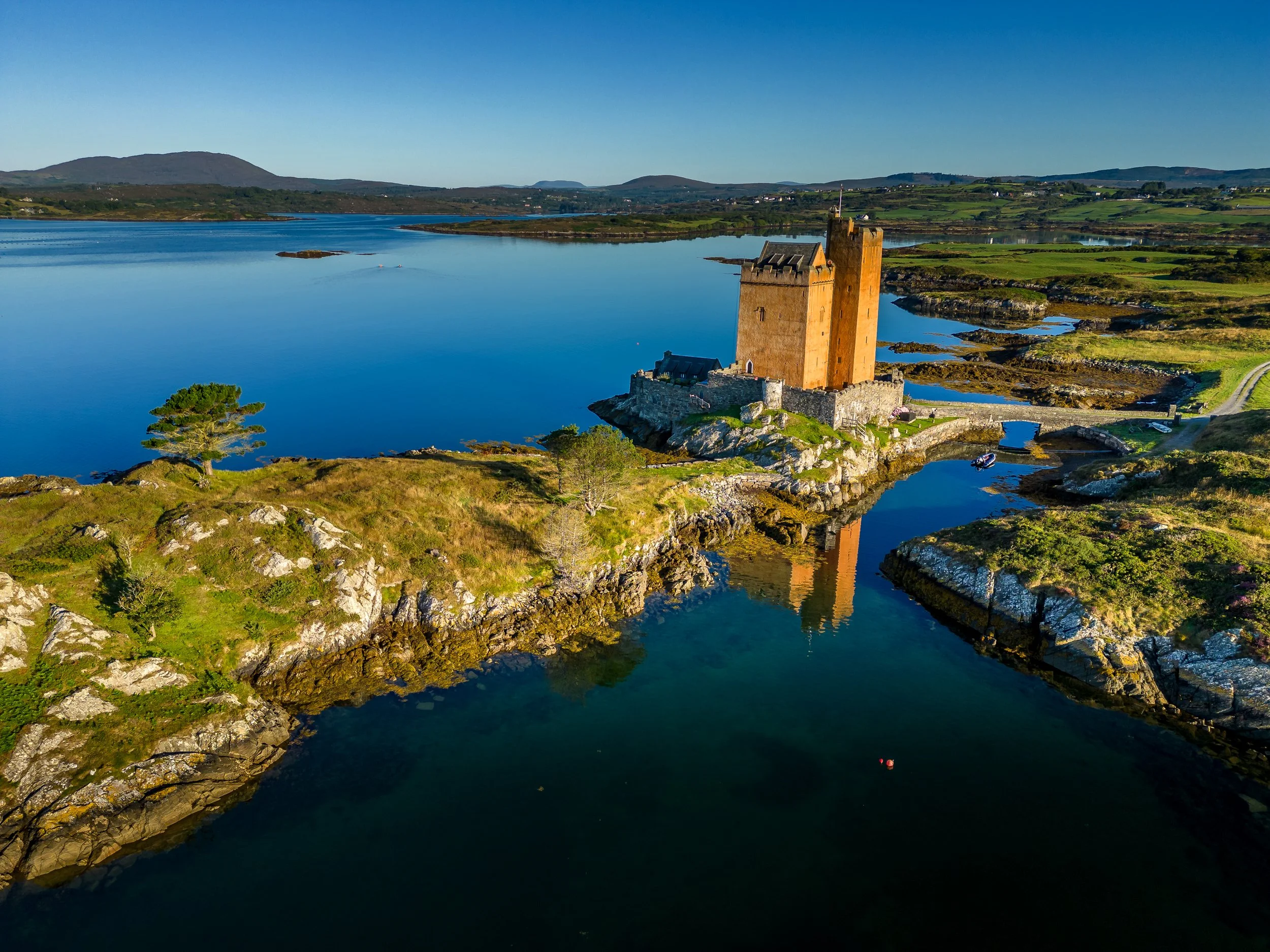 A castle on a small rocky island surrounded by calm water, with rolling green hills and mountains in the distance under a clear blue sky.