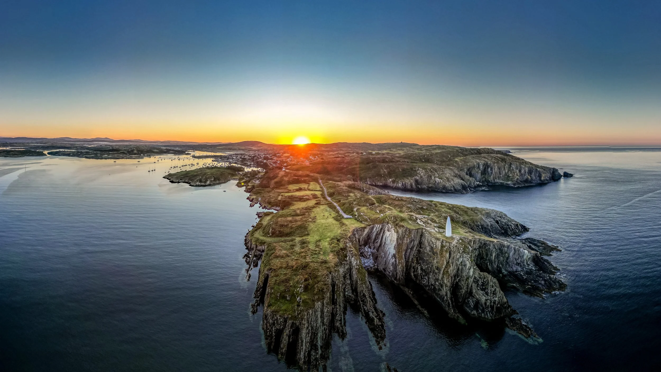 Aerial view of a coastal landscape at sunset, showing green grassy cliffs and a lighthouse on a point extending into the ocean.