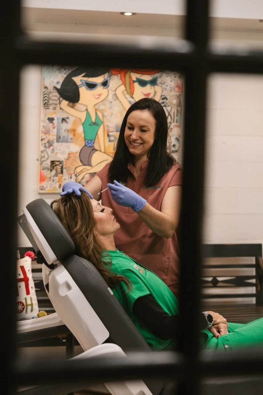 A woman receiving a cosmetic injection from a healthcare professional in a dental or medical office, seen through a window frame, with a colorful cartoon painting in the background.