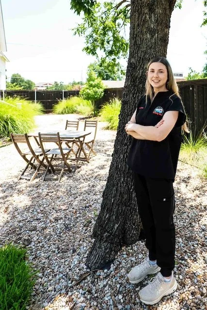 A young woman with long brown hair, wearing a black shirt and black pants, stands outdoors next to a large tree with her arms crossed and smiling. In the background, there is a patio with a wooden table and chairs, green grass, and trees.