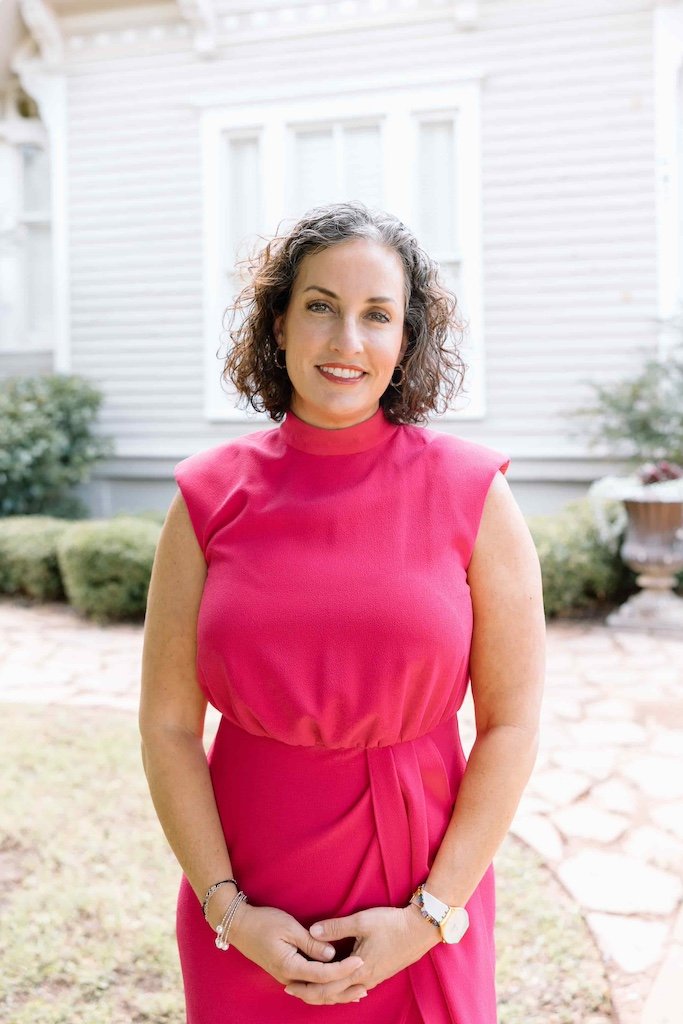 A woman in a bright pink sleeveless dress, smiling, standing outdoors in front of a white house with shrubs.
