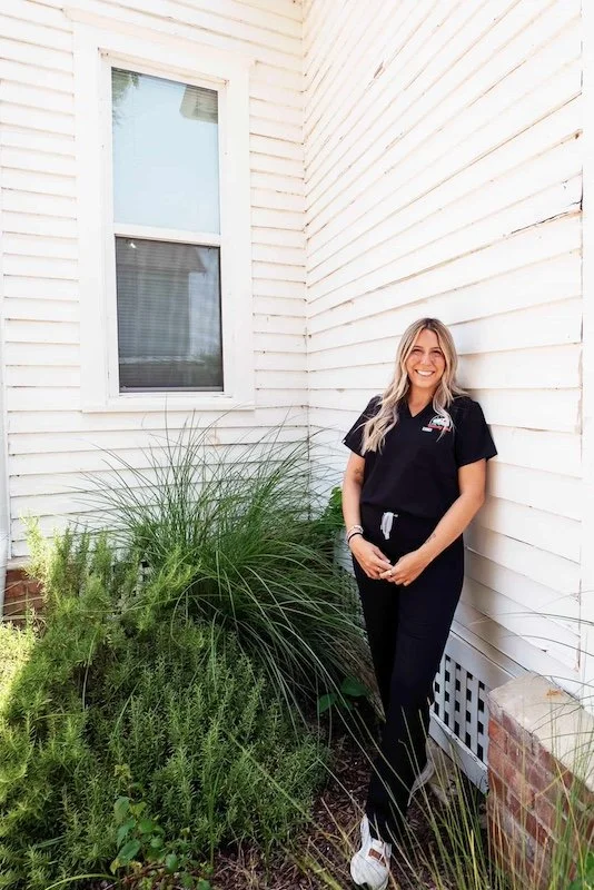 A woman standing next to a green plant beside a white house wall, smiling, wearing black scrubs and white sneakers.