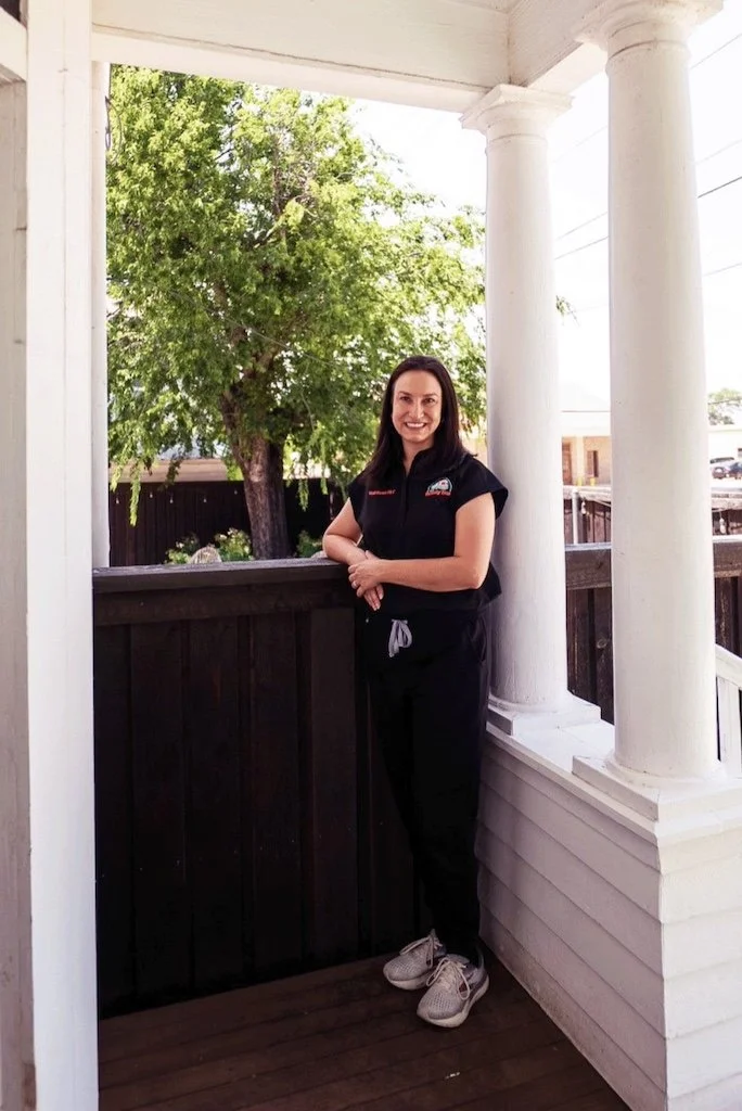 A woman standing on a porch with white columns, a dark wooden railing, and a tree in the background, smiling at the camera.