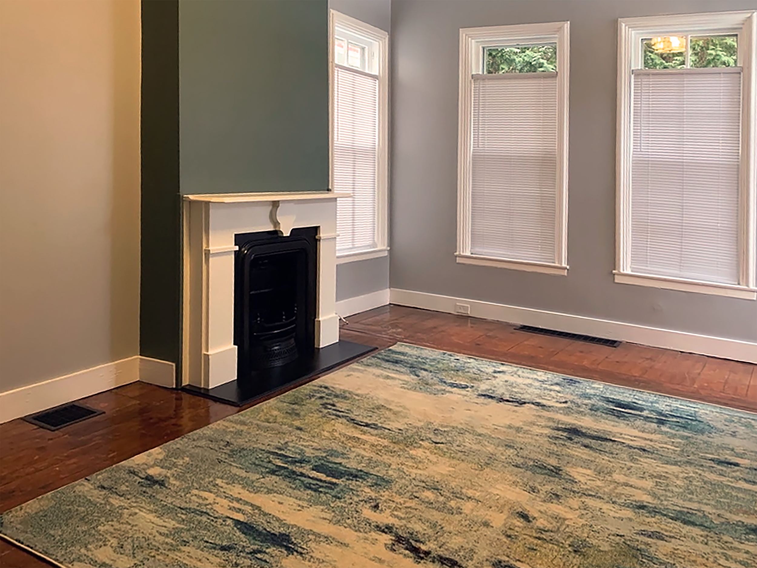 Empty living room with hardwood floors, a colorful area rug, three large windows with blinds, a white fireplace, and light blue walls.