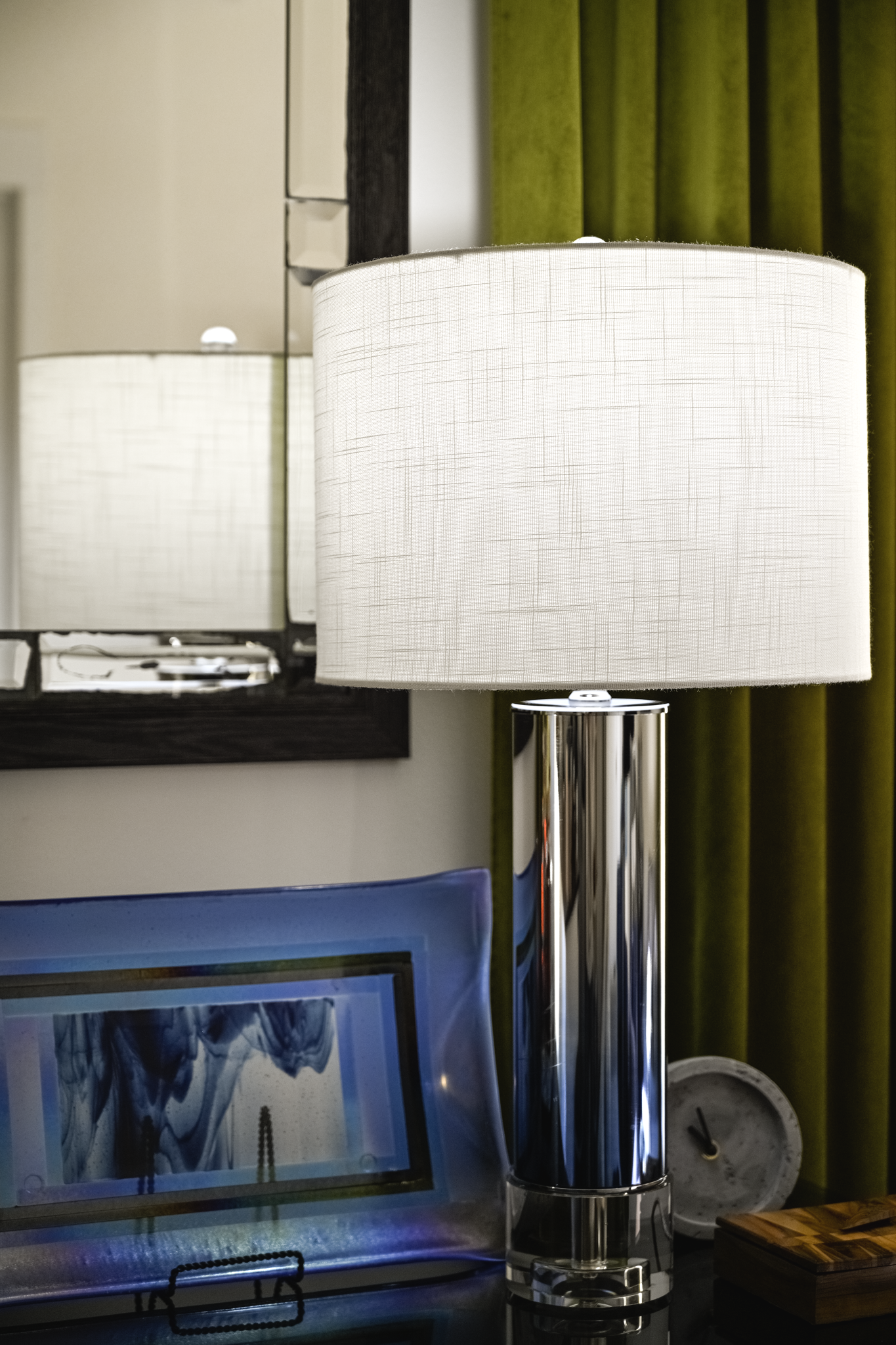 A modern table lamp with a white shade and shiny chrome base on a dark surface, with a mirror, framed artwork, and a white clock in the background.