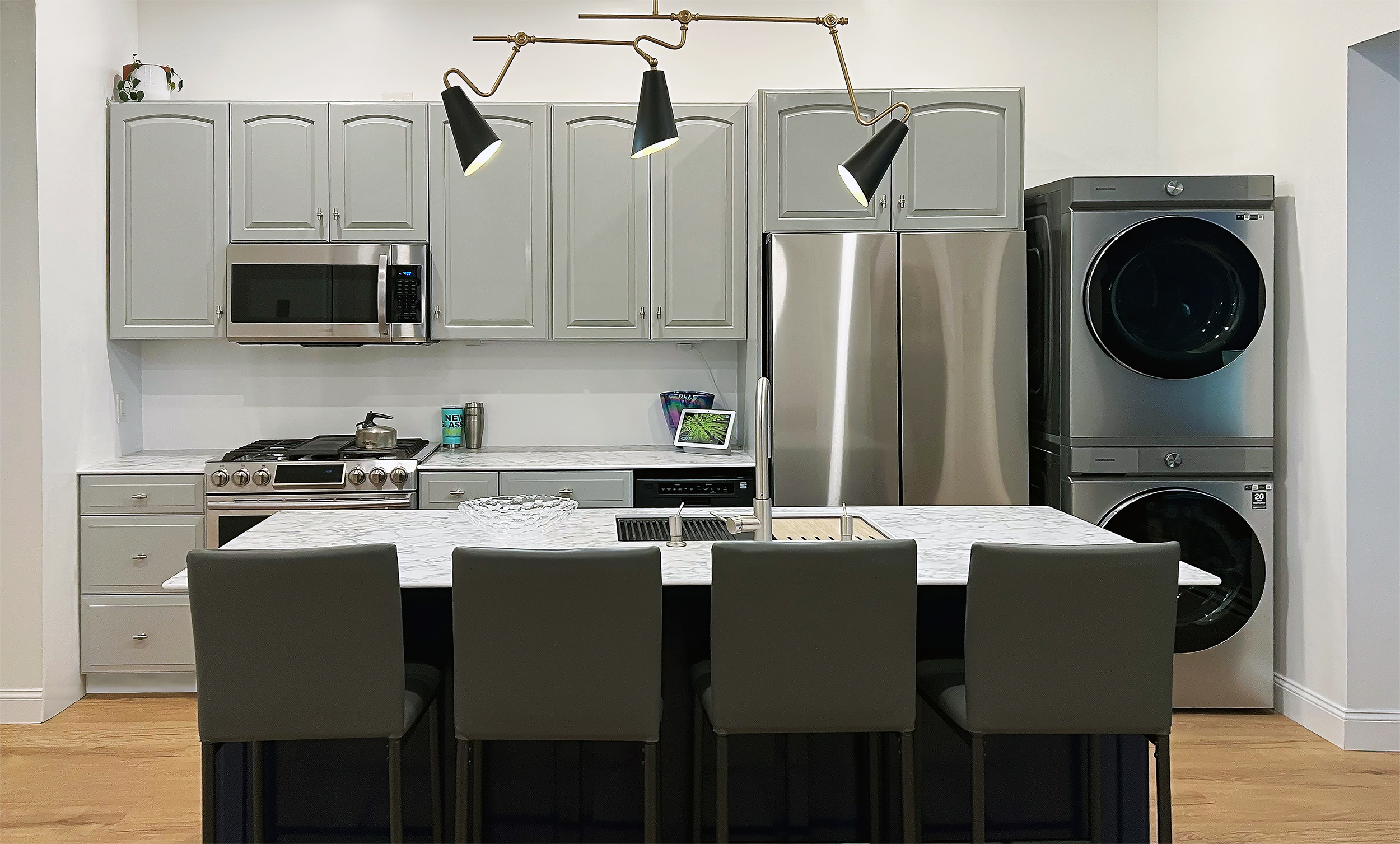 Modern kitchen with gray cabinets, stainless steel appliances, and a marble countertop island with four chairs, overhead black and gold pendant lights, laundry machines, and a microwave.
