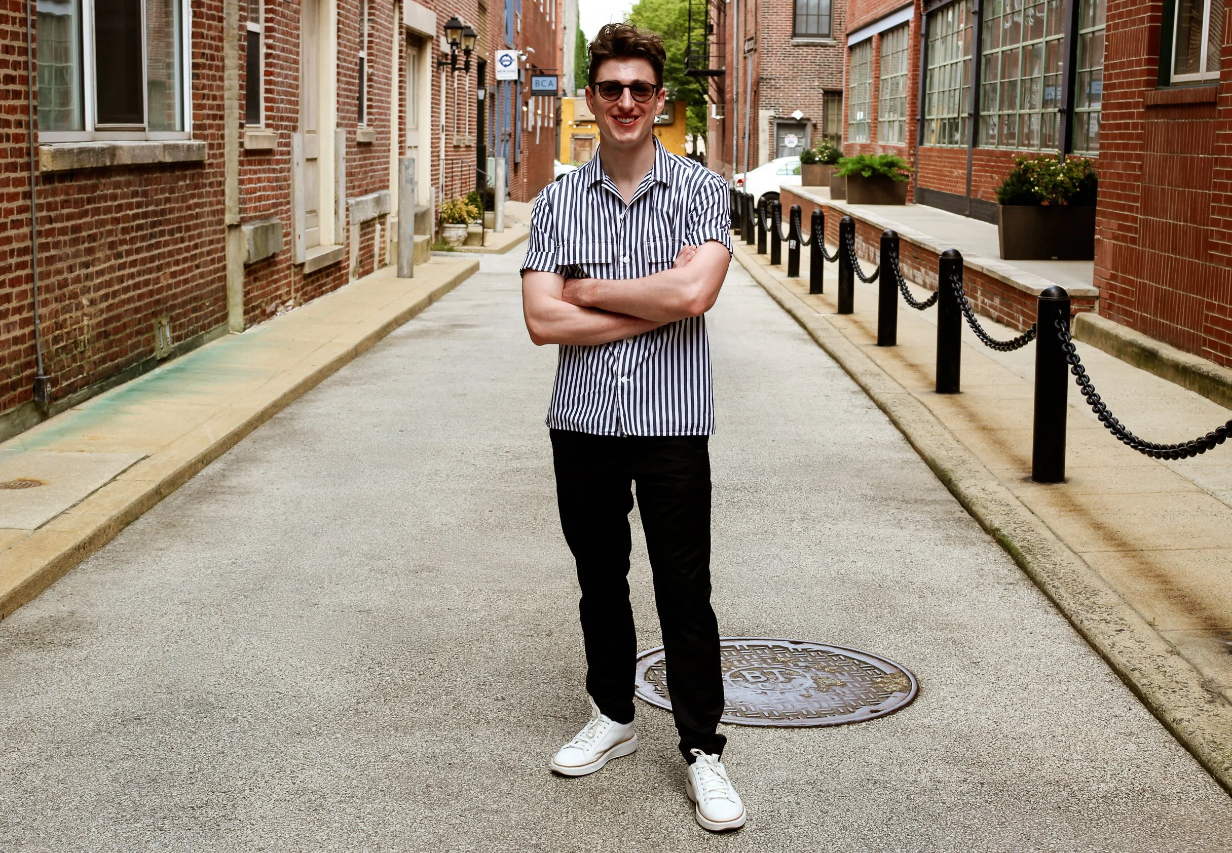 A young man standing with arms crossed, smiling, on a quiet city street lined with brick buildings, with potted plants and black chain-link barriers.