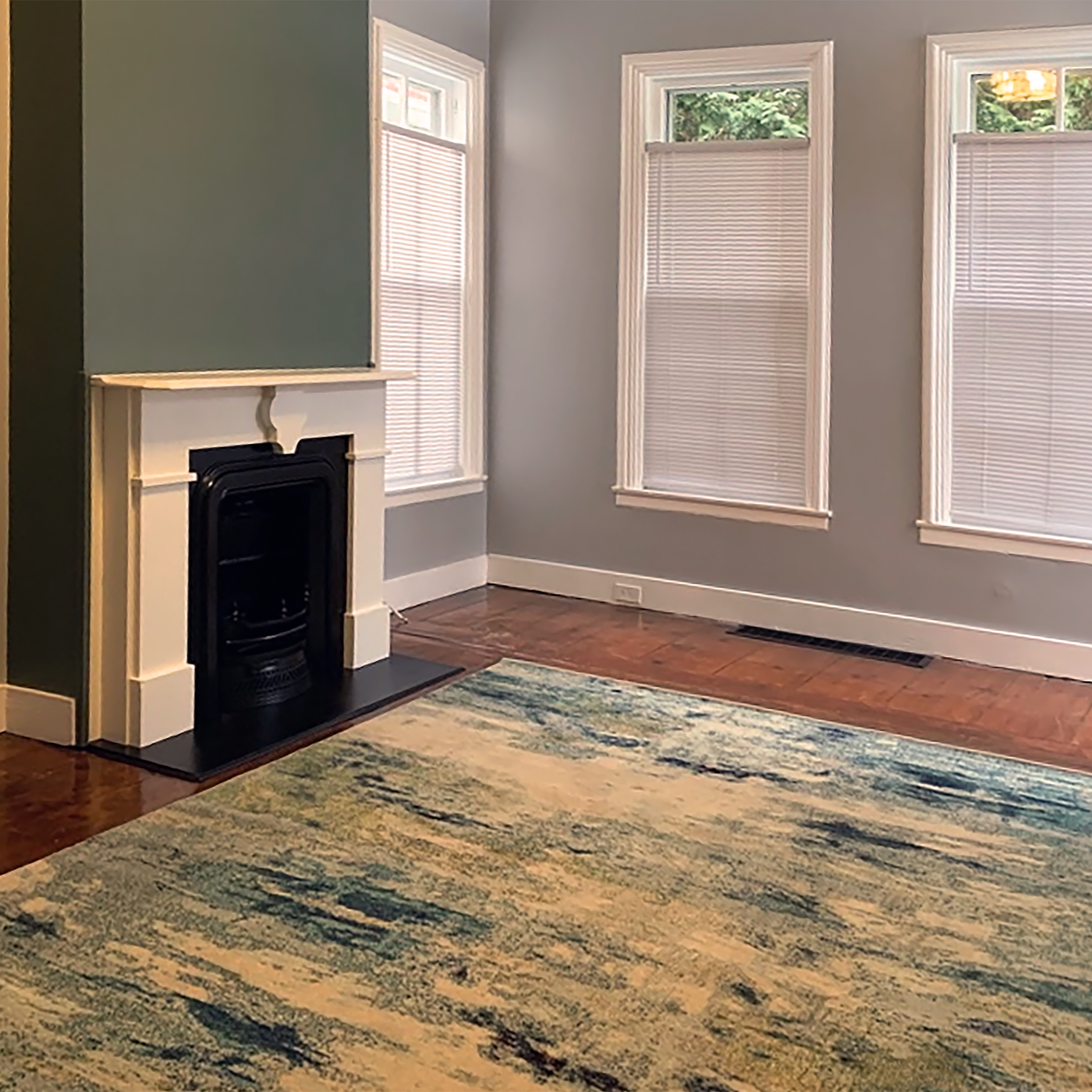 Empty living room with hardwood floors, a colorful area rug, three large windows with blinds, a white fireplace, and light blue walls.