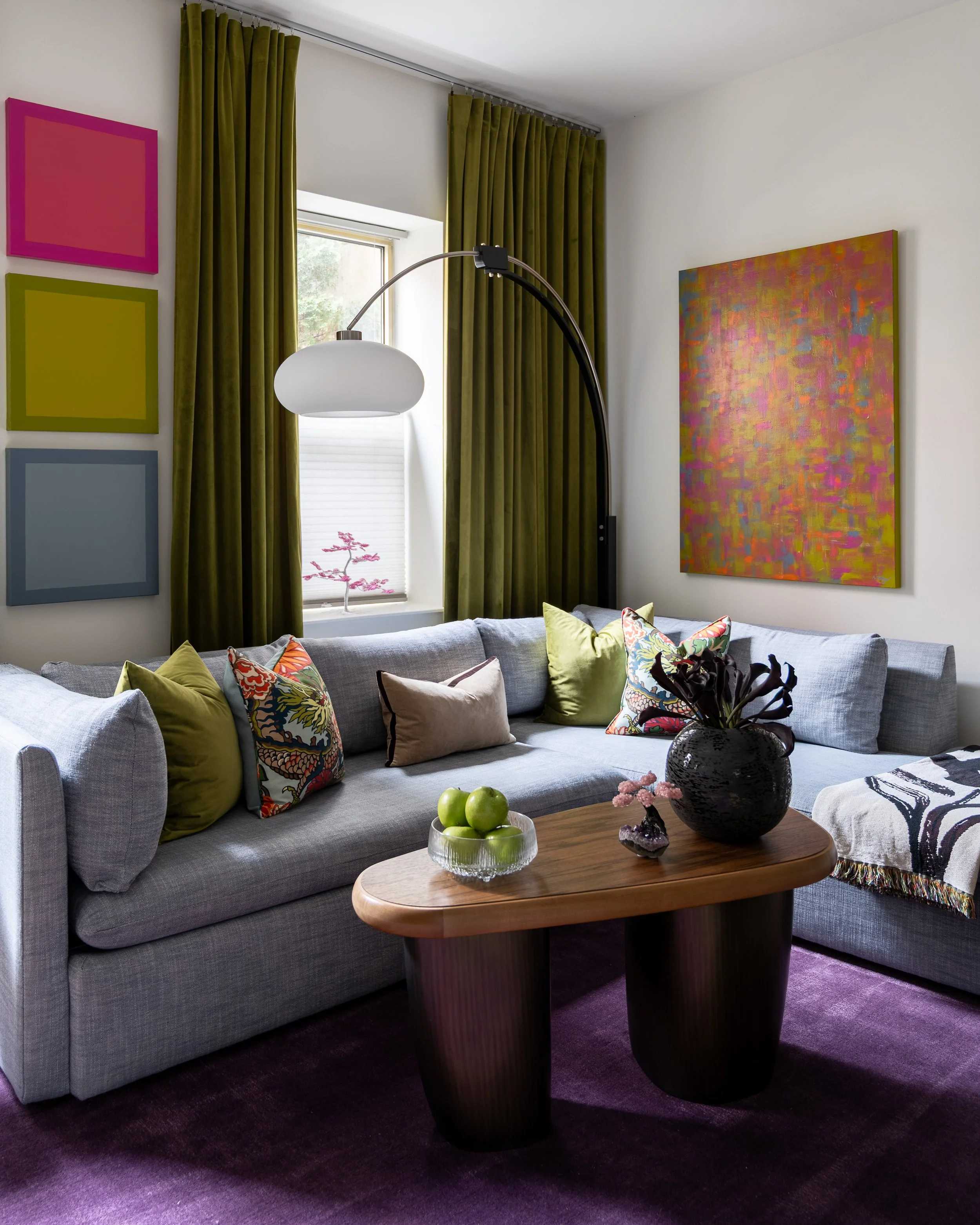Eclectic Philadelphia living room design with olive green curtains, colorful art, and gray sectional.