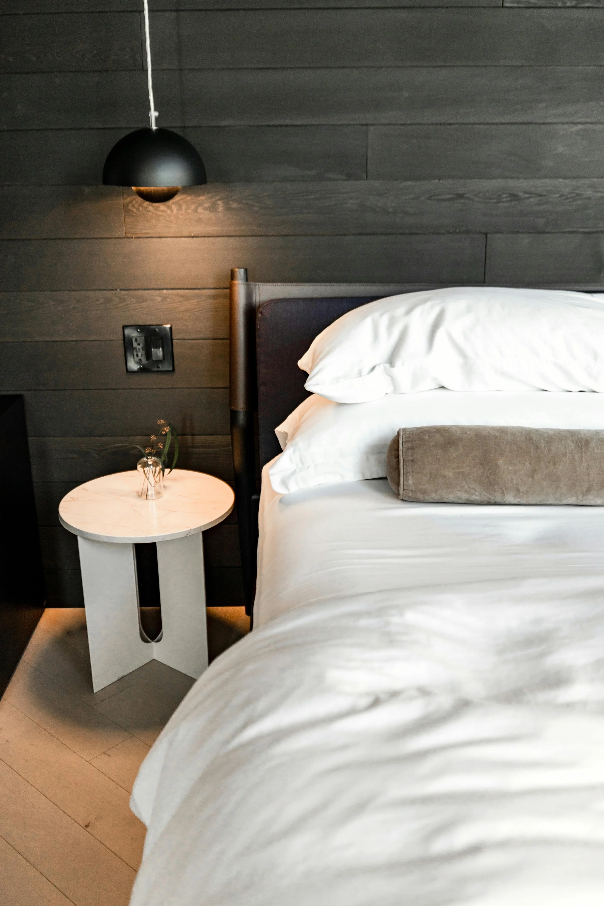 Close-up of a neatly made bed with white bedding and a cylindrical pillow, in a modern bedroom with dark wood wall panels and a small round bedside table with a small vase and flower. A black pendant light hangs above the bed.