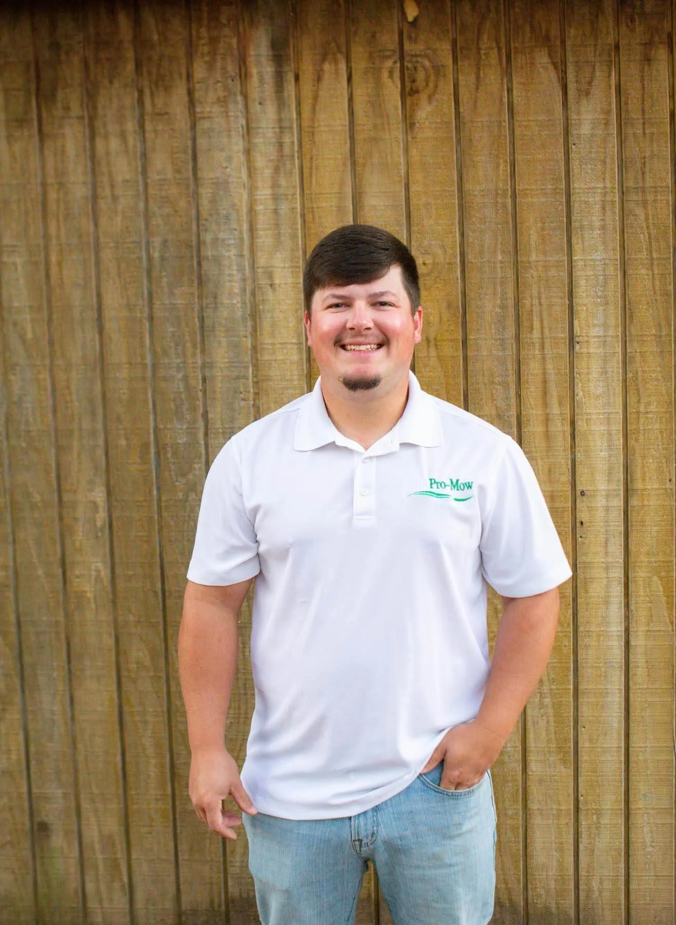 Garrett Bowers Headshot, wearing a white polo shirt with 'Pro-Mow' embroidered on it, standing in front of a wooden fence with his right hand in his pocket.