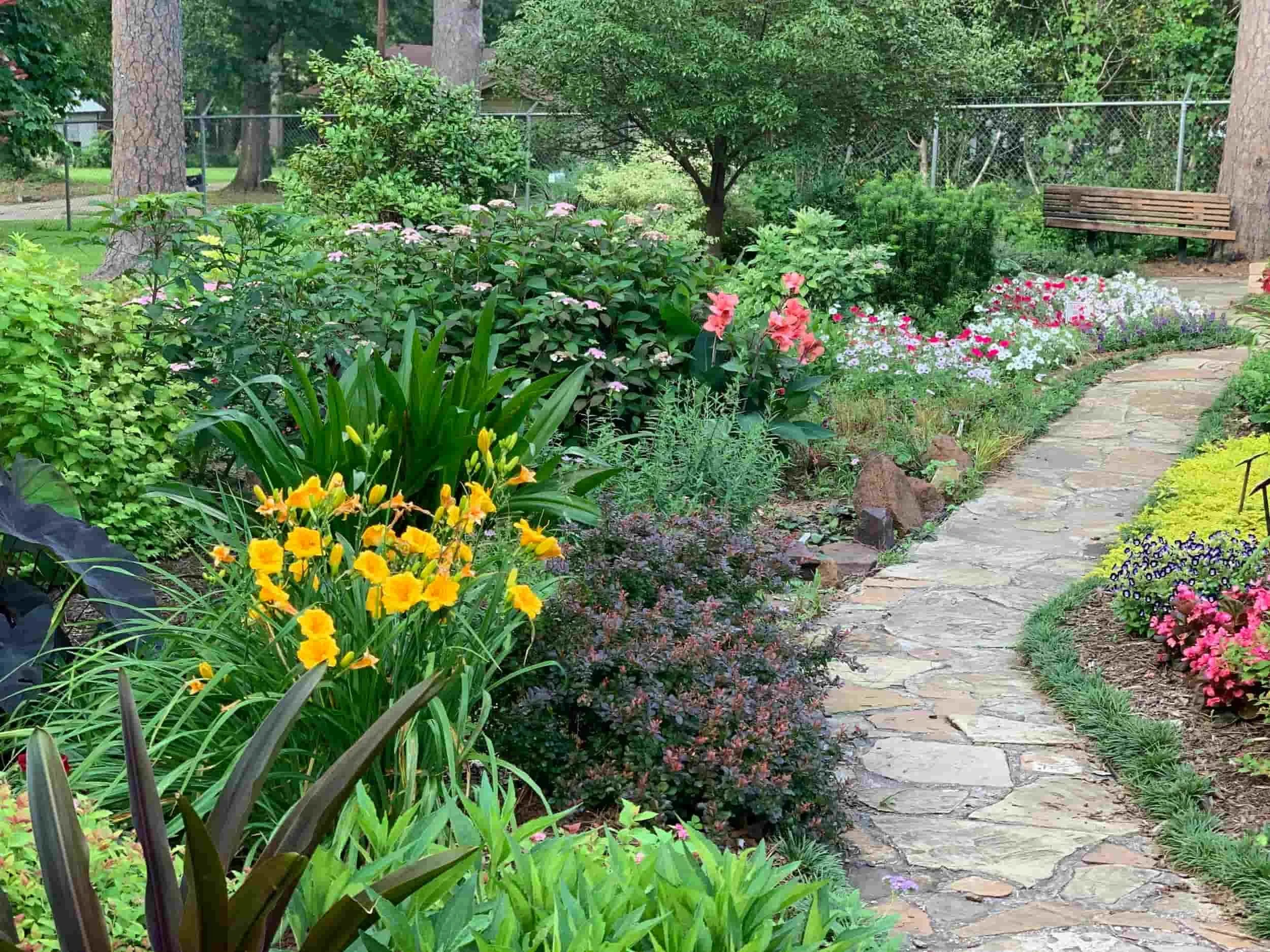 A garden scene with a stone pathway and harscaping by promow lawncare surrounded by colorful flowers and lush green foliage, with a wooden bench in the background.