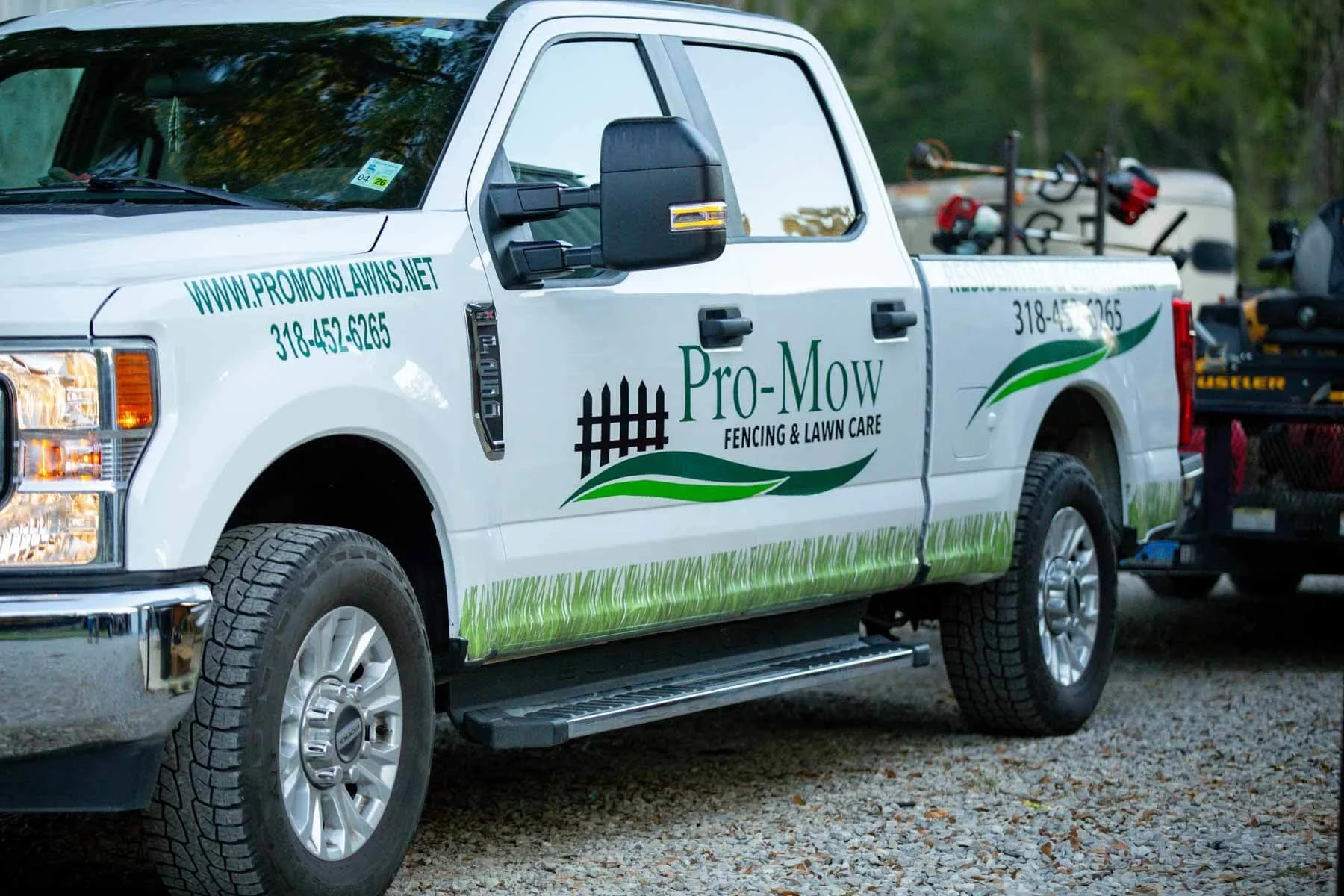 A white pickup truck with Pro-Mow Fencing & Lawn Care branding, parked on a gravel surface, with a utility trailer attached carrying lawn equipment and tools.