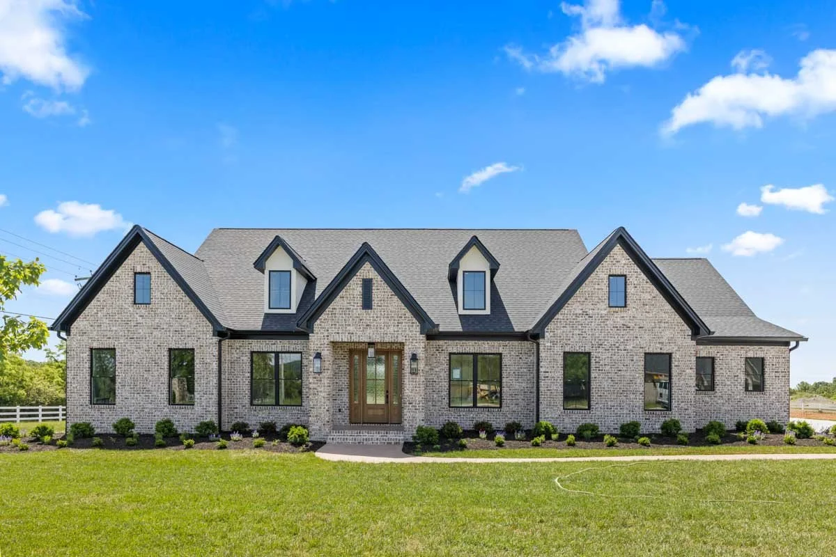 A large brick house with multiple gabled roofs, surrounded by a green lawn and small bushes, under a bright blue sky with a few clouds.