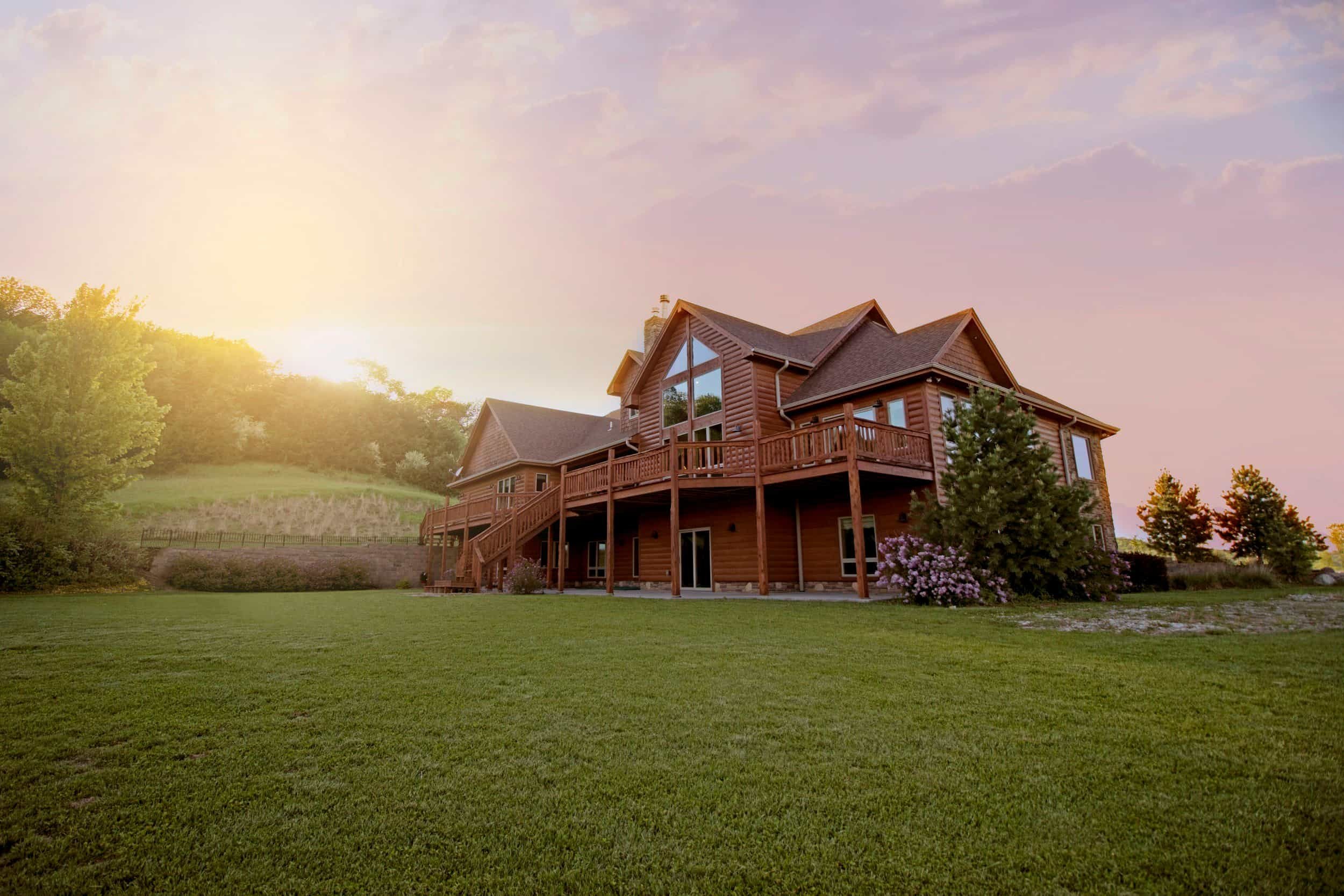 A house with a well-maintained lawn and large windows and a multi-gabled roof. The background includes a hillside with trees under a sunset sky with pink and purple clouds.