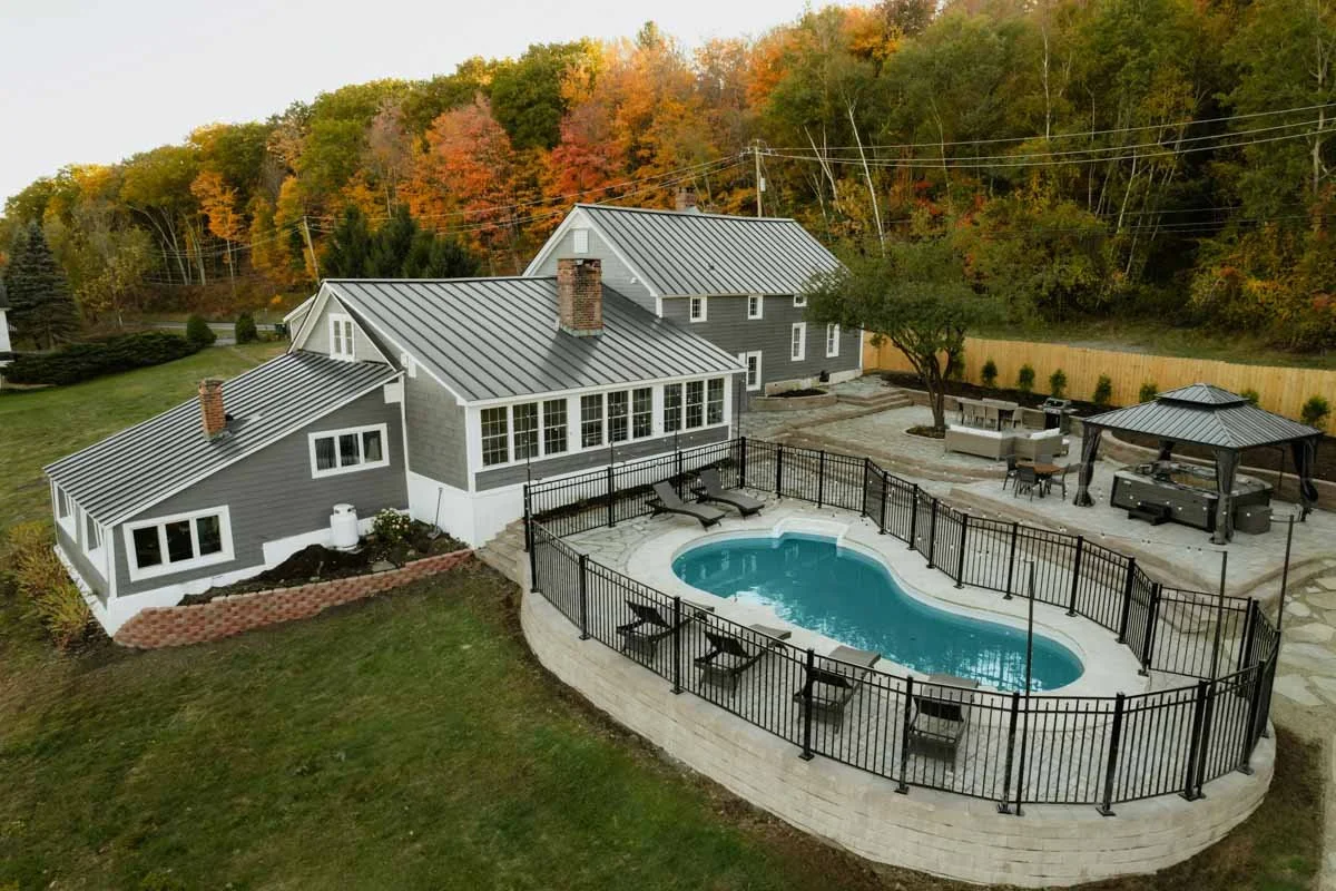 A pool and patio by a large gray house with a metal roof, surrounded by a lawn and a metal fence.