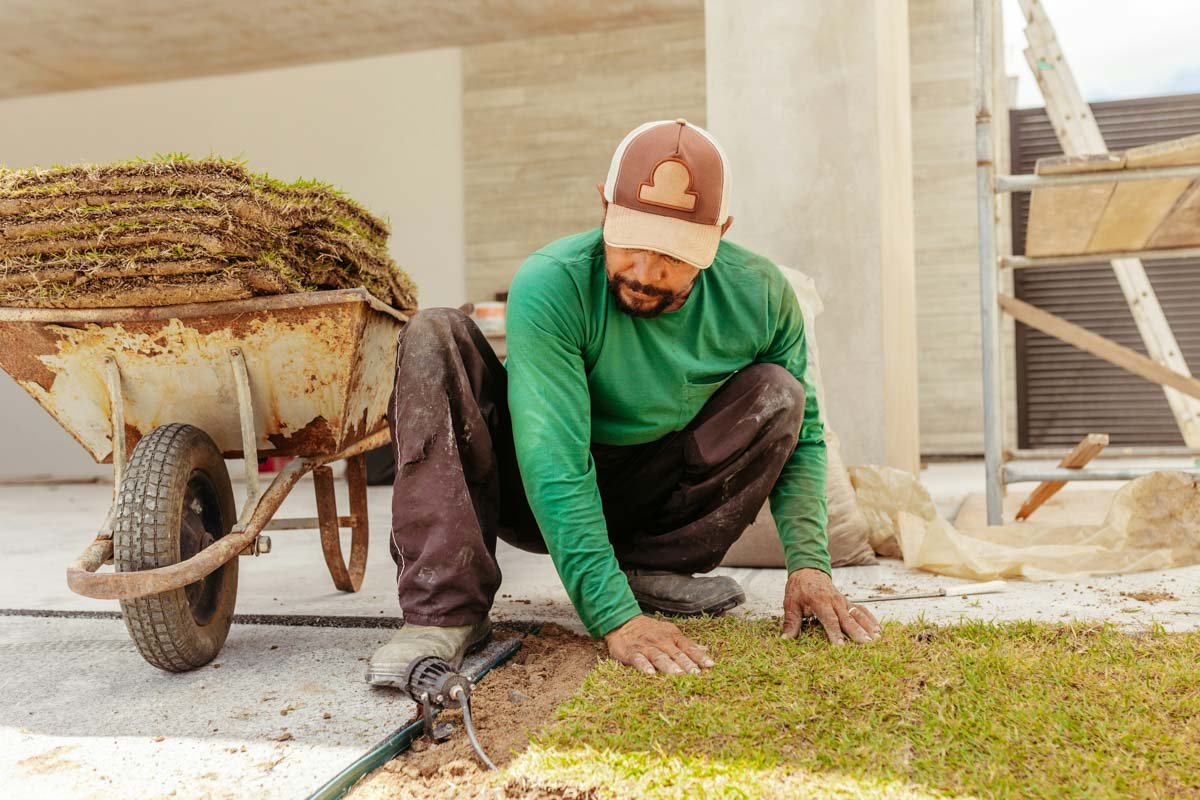A man wearing a green shirt kneels on the ground, working on installing grass in a yard with building materials and a wheel-borrow around him. Behind him is a stone house and gate.