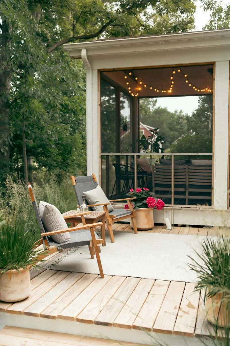 A patio with chairs, potted plants, pink flowers, and a screened porch with string lights behind.