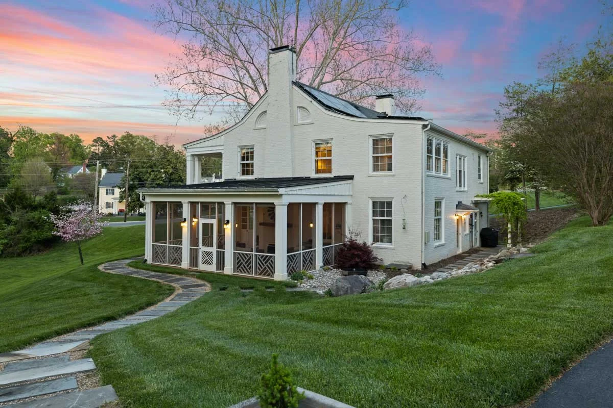 A white house with a screened porch, a curved roof, trees and a sunset sky in the background.